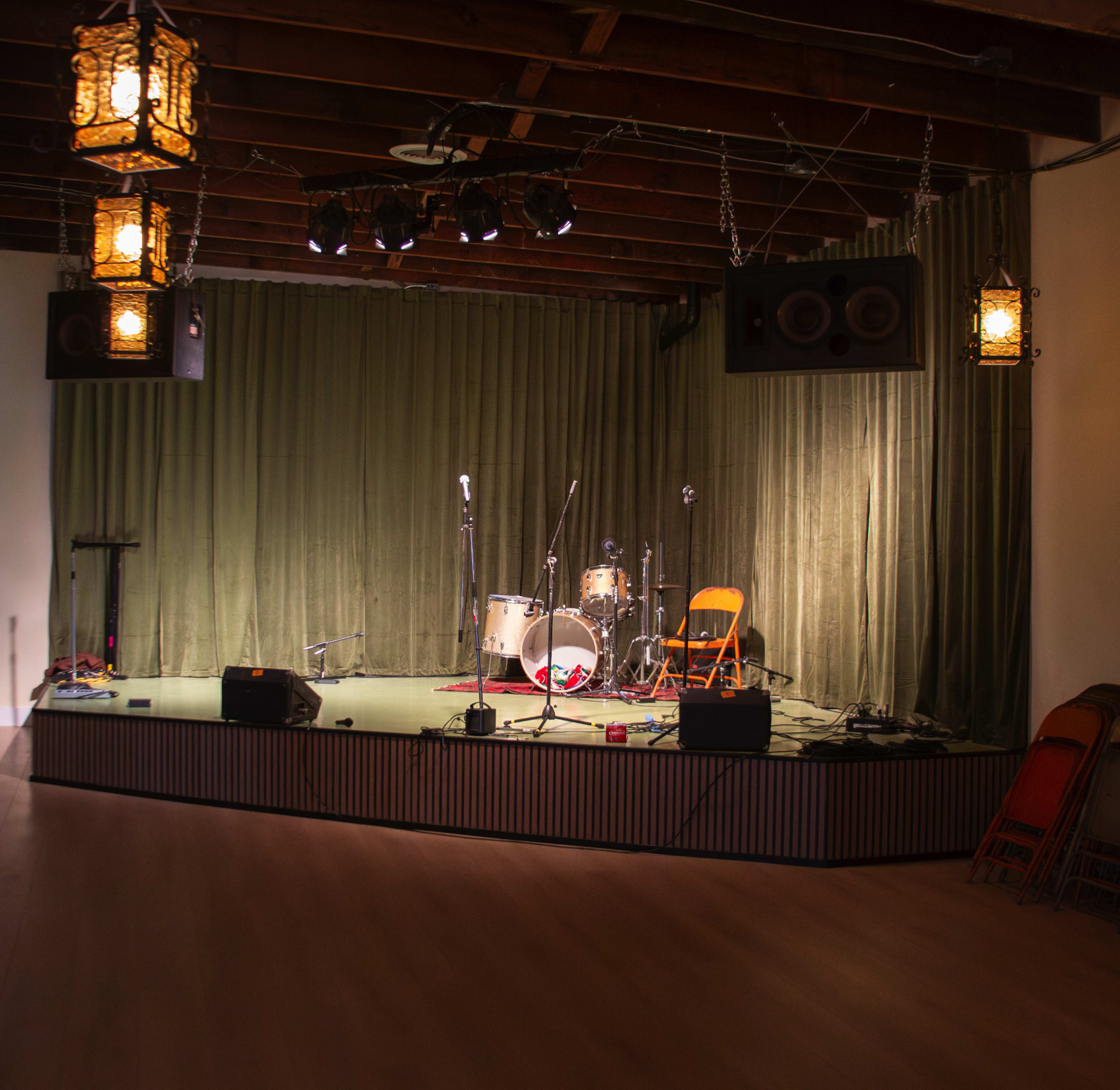 A small stage is set up with musical equipment and a single orange chair, surrounded by green curtains and overhead lanterns.