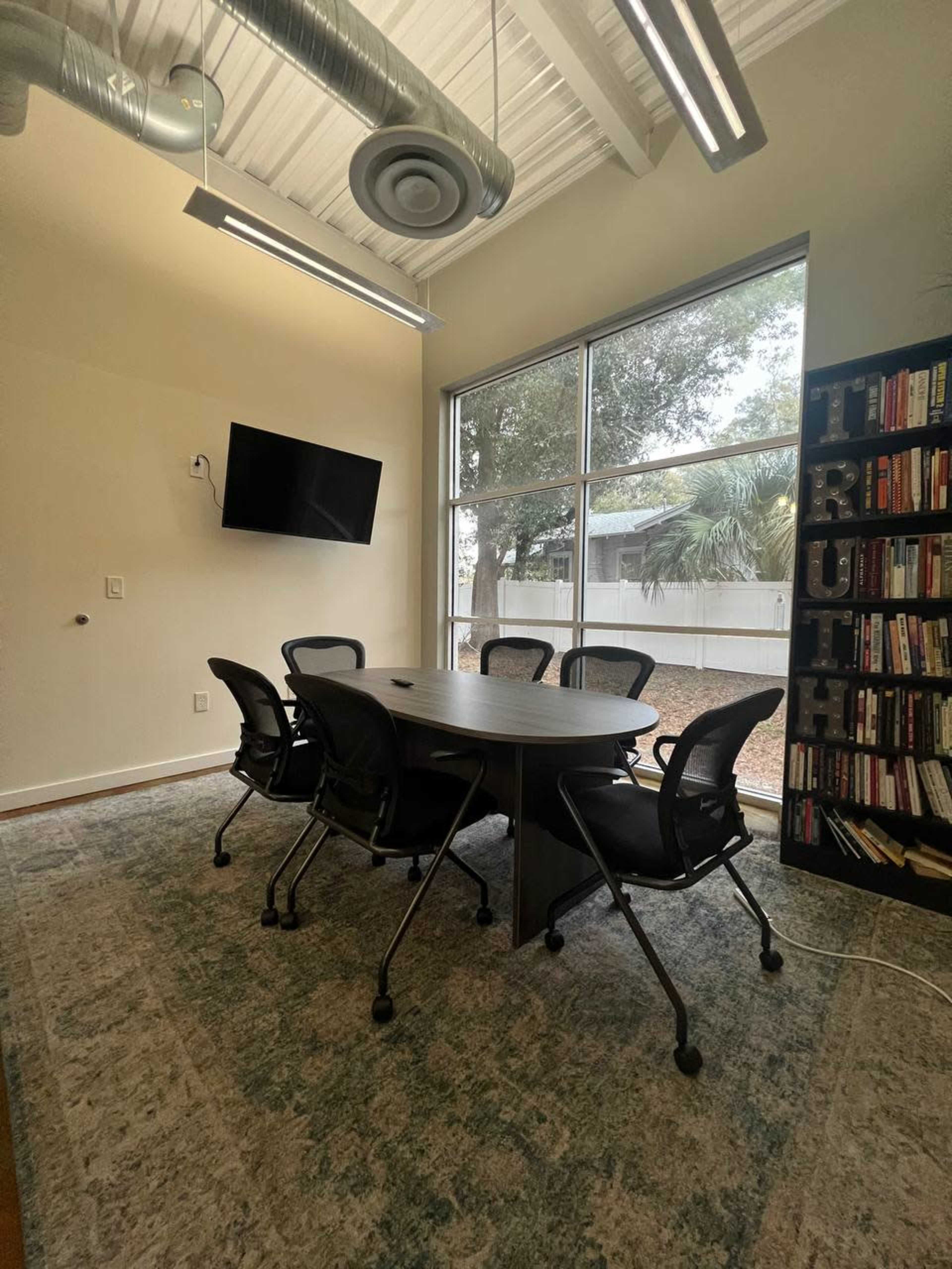 A conference room features a round table with six chairs, a wall-mounted television, and a bookshelf filled with books, all under a bright window.