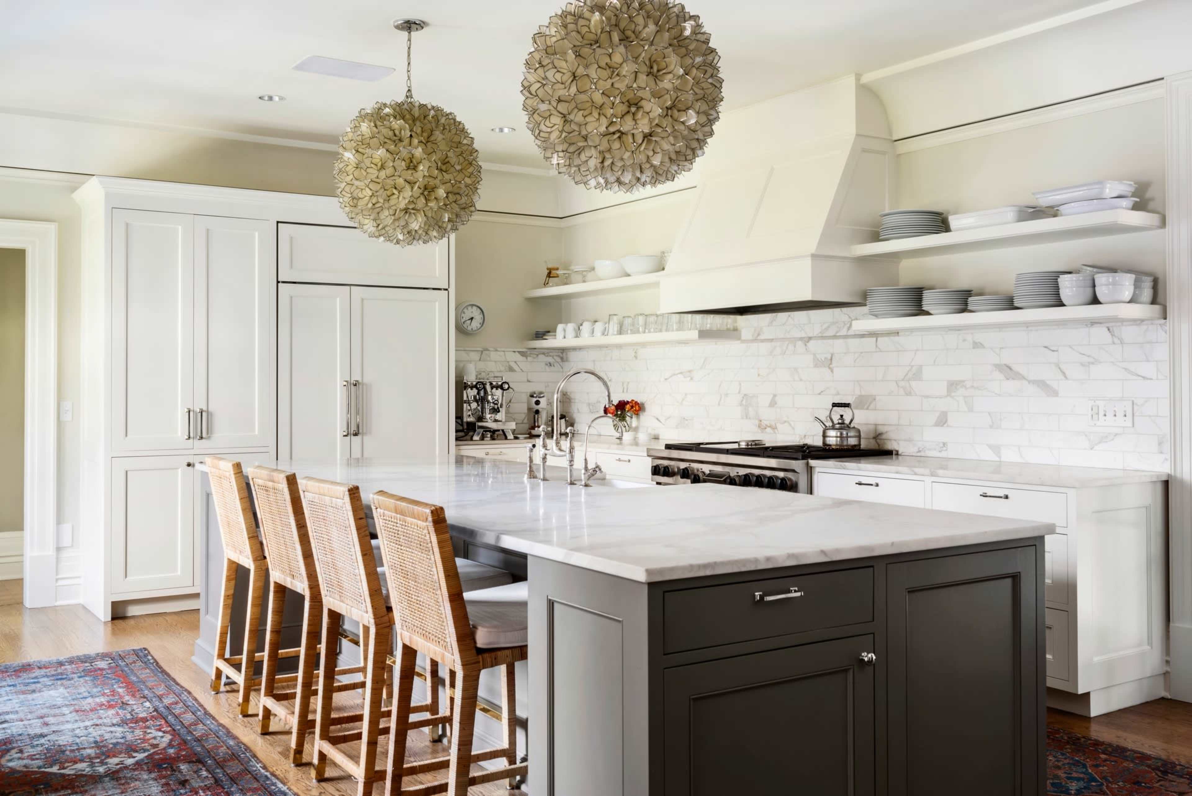 A modern kitchen featuring a large island with four wicker bar stools, marble countertops, and white cabinetry, with decorative spherical light fixtures overhead.