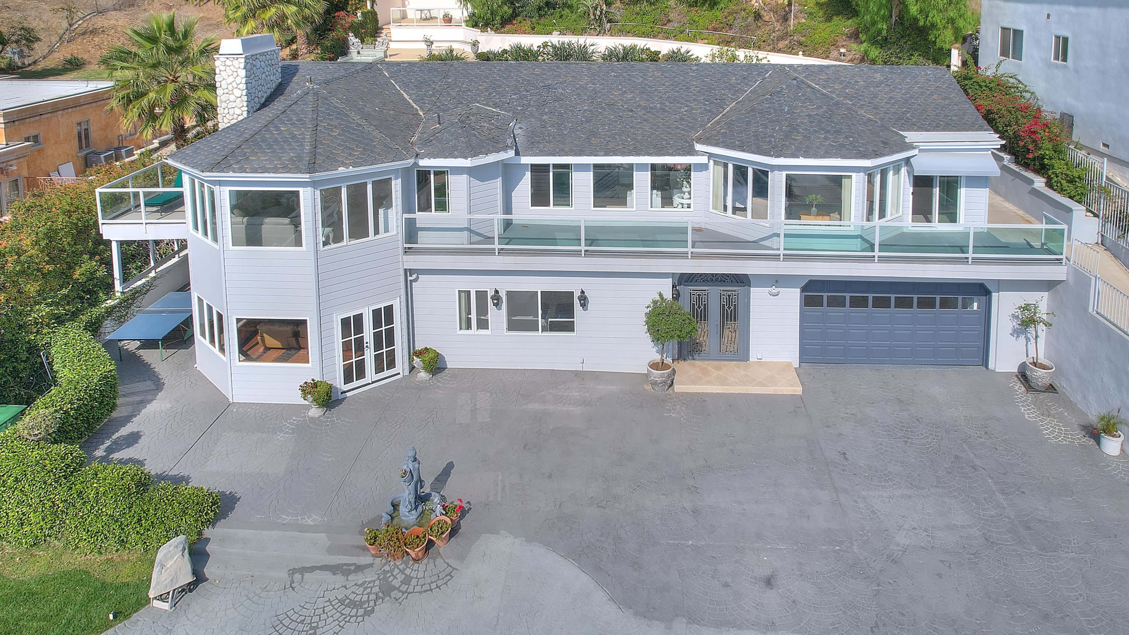 A large, modern two-story house with a gray exterior, multiple large windows, and a balcony, situated on a landscaped property with a driveway and decorative planters.