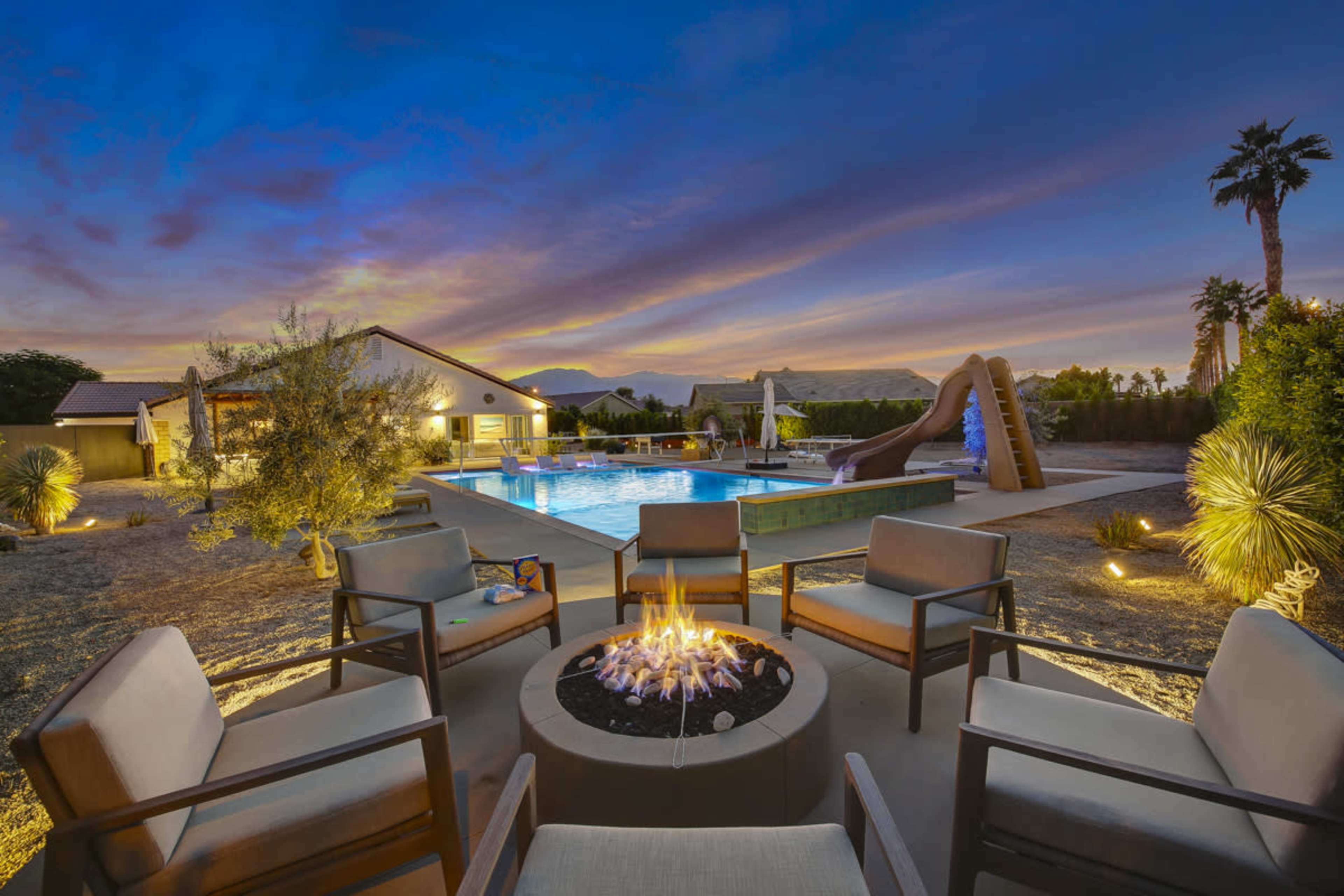 A circular fire pit with chairs surrounds a pool area that features a water slide and illuminated landscaping against a twilight sky with mountains in the background.