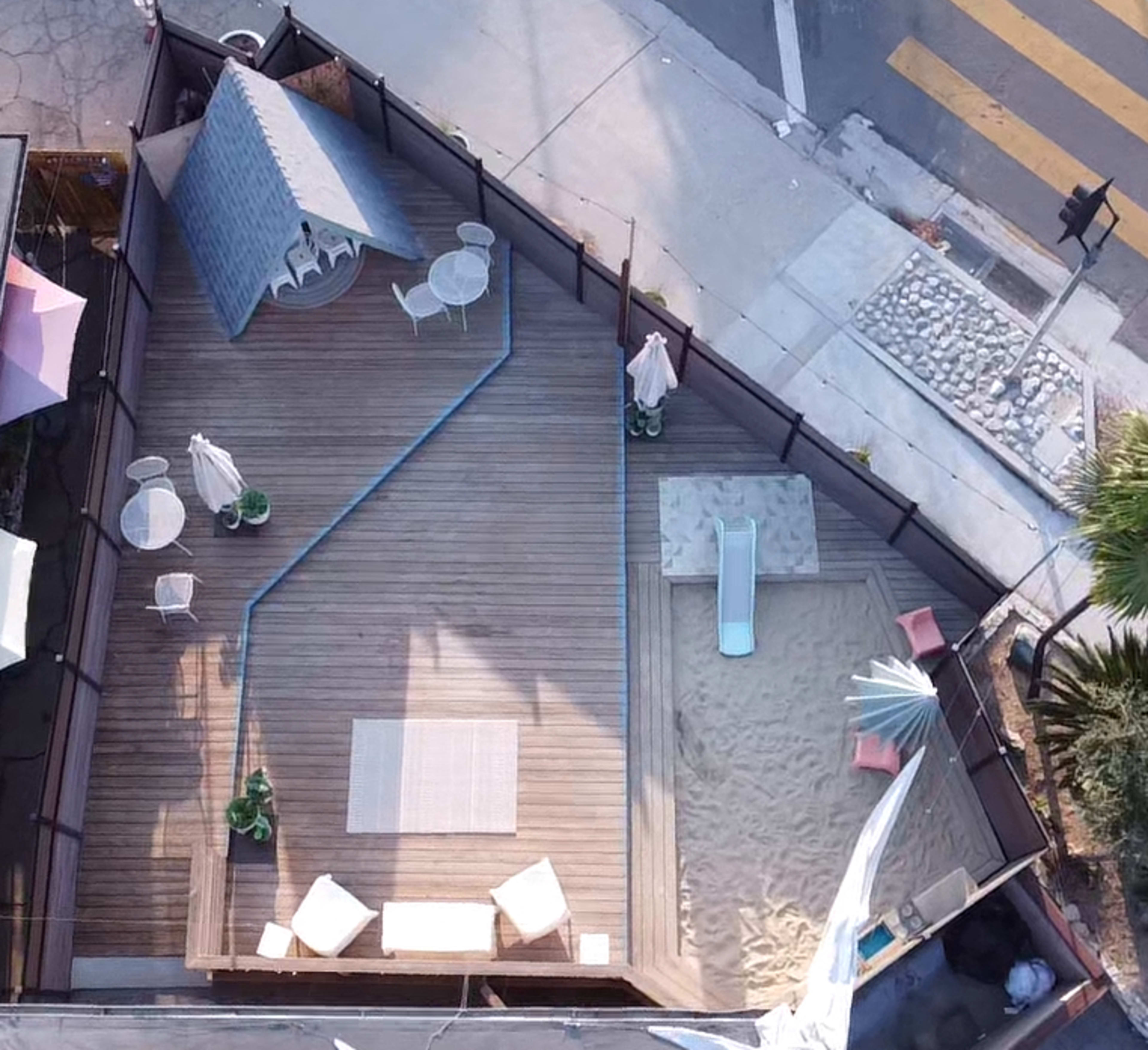 An aerial view of a wooden deck area with lounge furniture, a small pool, and a sand play area, all enclosed by a fence near a street intersection.