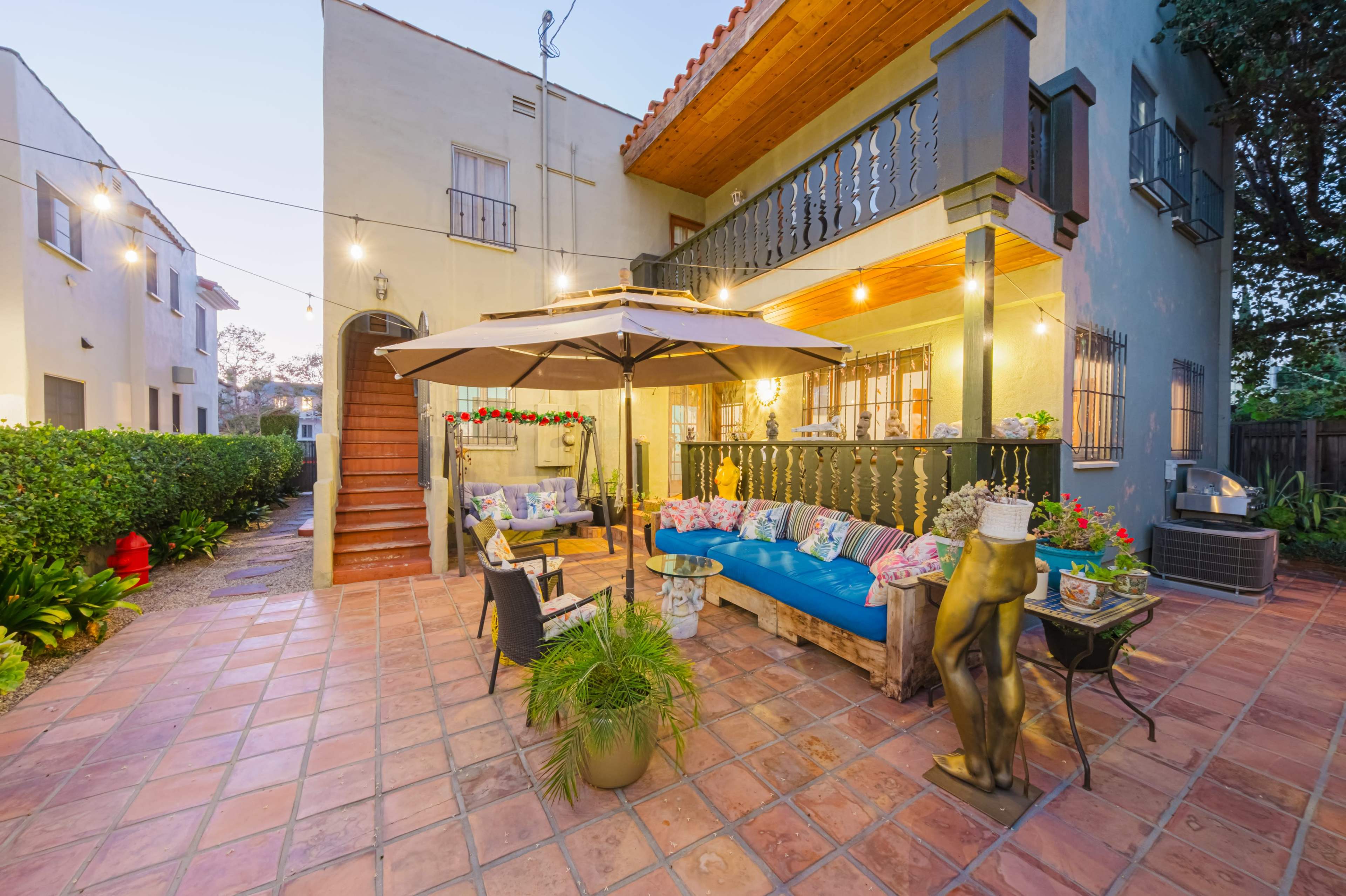 The image shows a colorful outdoor seating area with a blue couch, surrounded by plants and decorative elements, under a canopy near a two-story building with string lights illuminating the space.