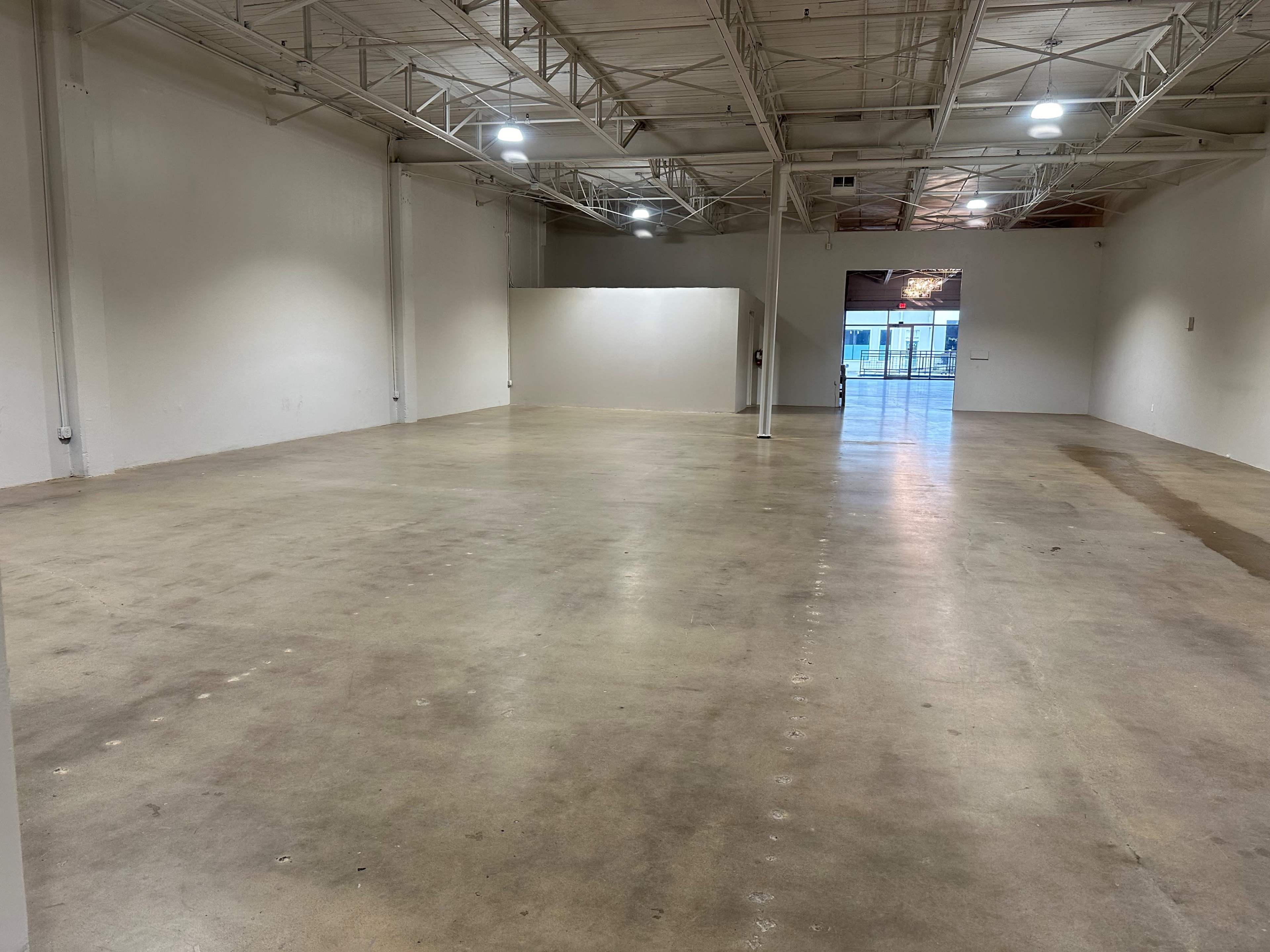 An empty industrial space with concrete flooring and white walls, illuminated by ceiling lights.