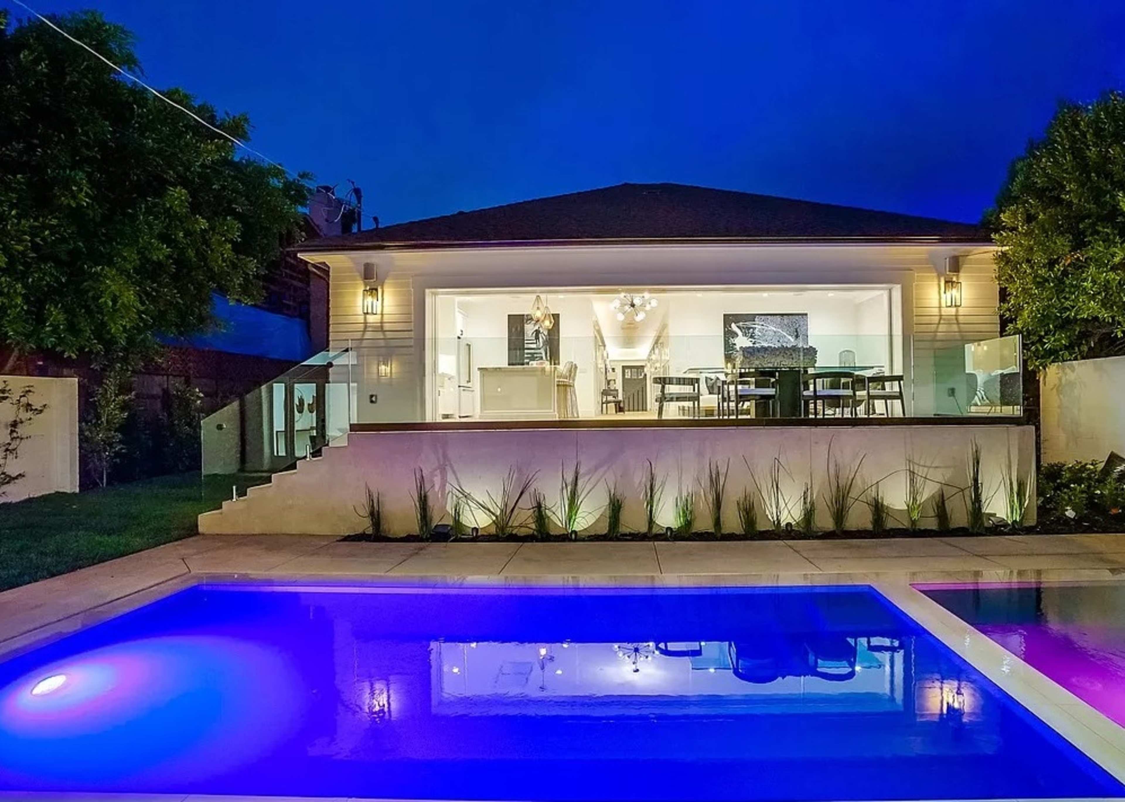 A modern house with large glass doors overlooks a lighted swimming pool at dusk.