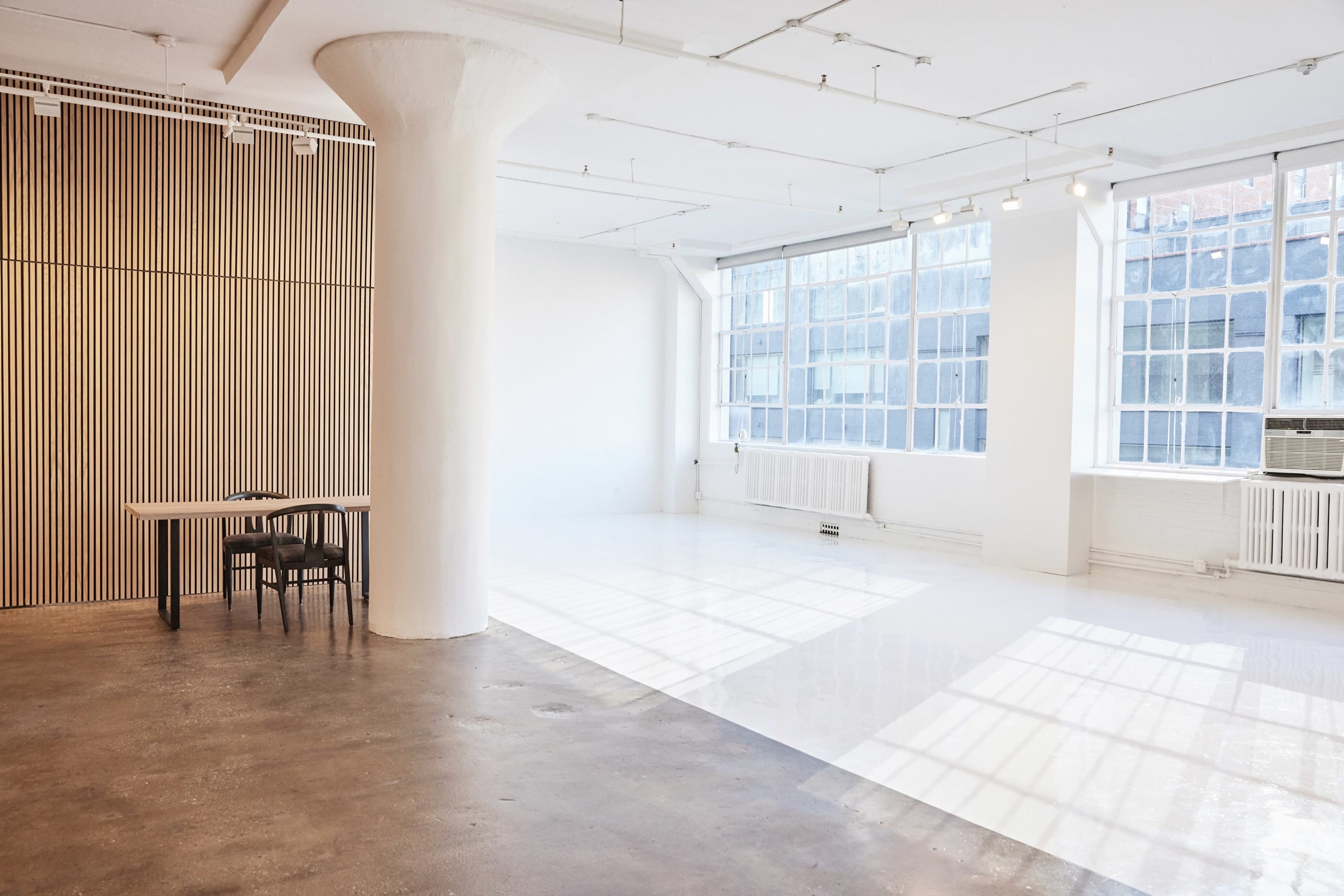 A spacious, well-lit indoor area with large windows, a table, and a few chairs against a wall with vertical wooden paneling.