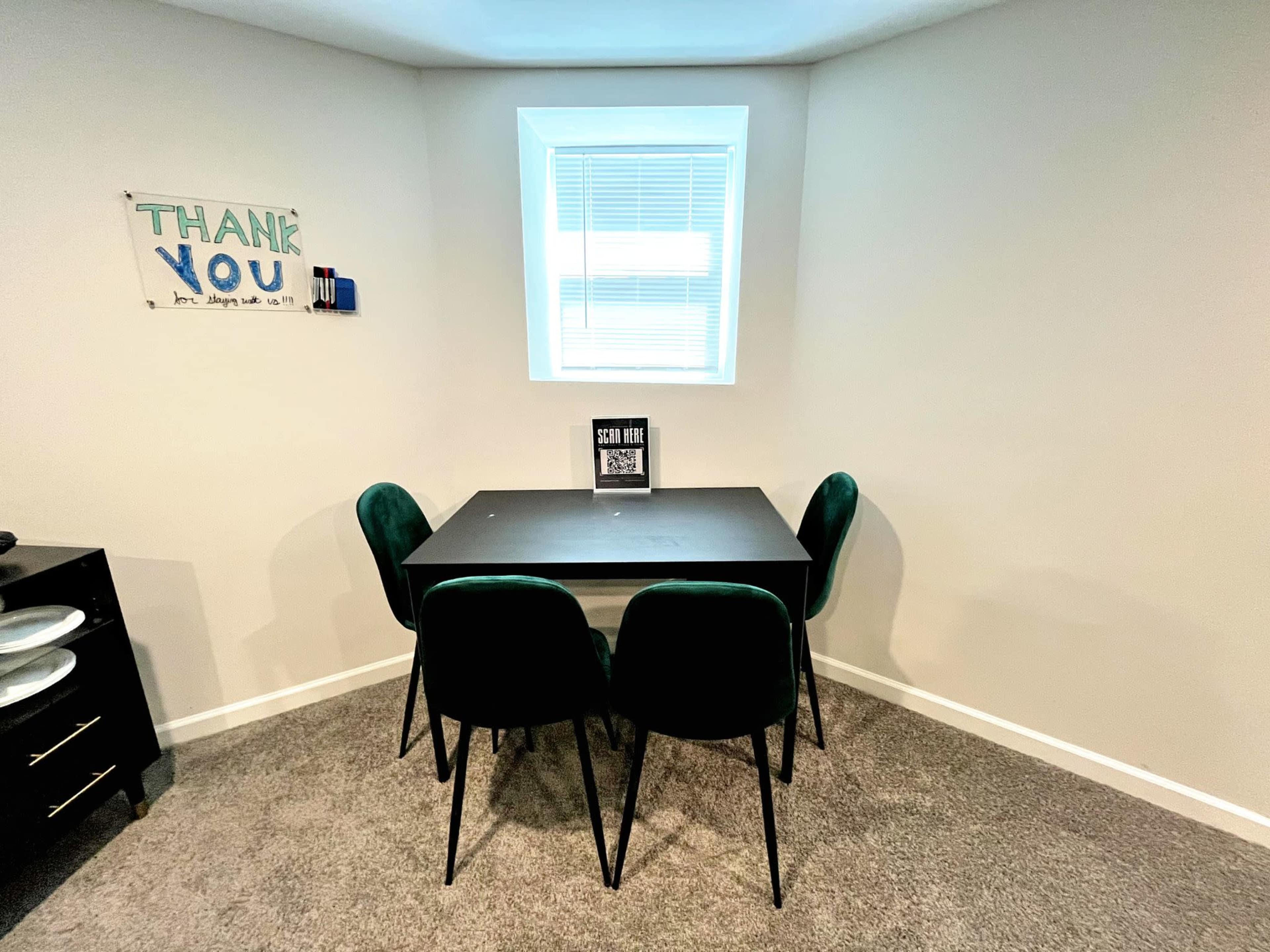 A black table surrounded by four green chairs is positioned in a corner of a room with a window and a "Thank You" sign on the wall.