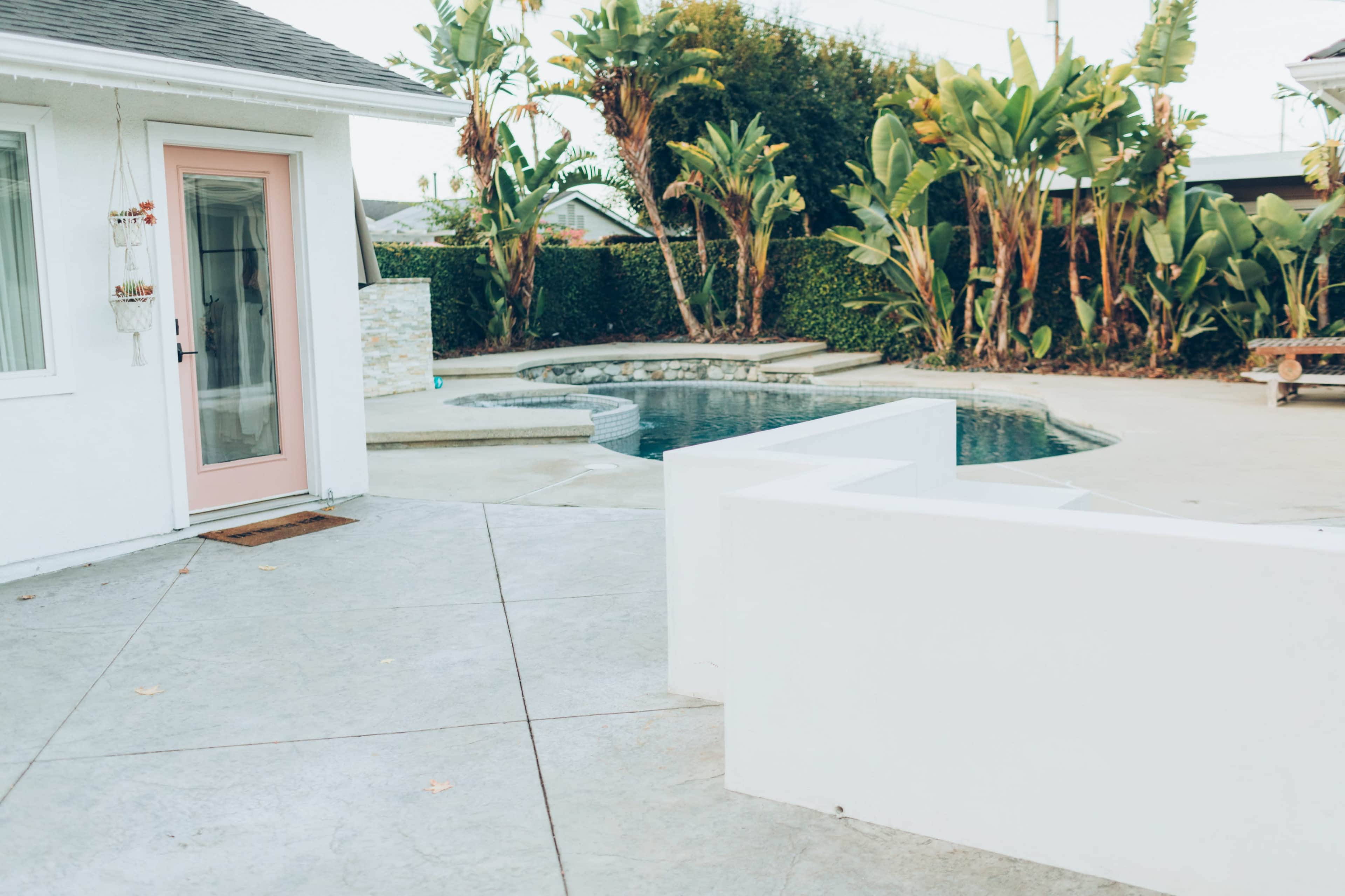 A backyard with a pool surrounded by palm trees and a white structure with a pink door.