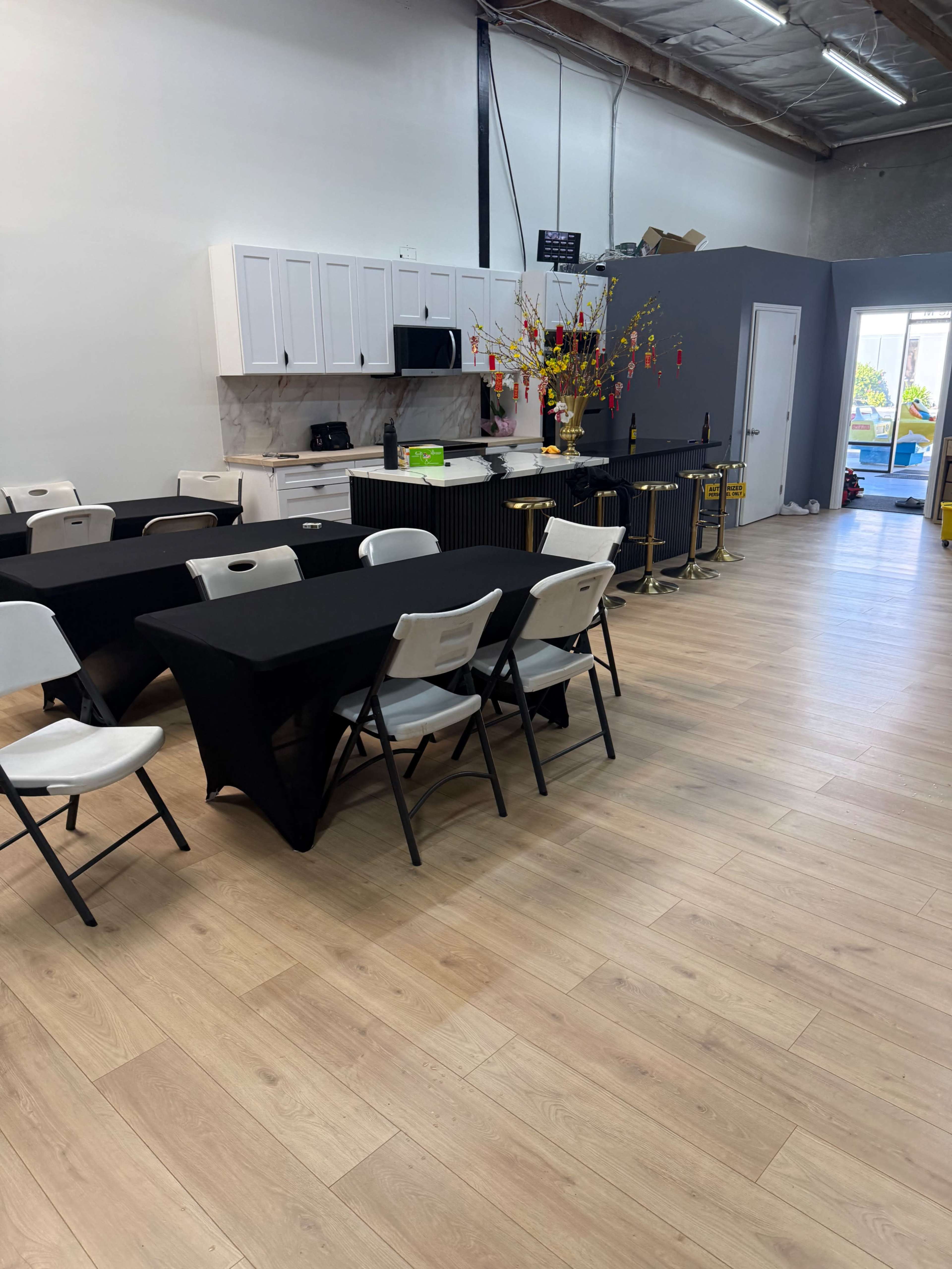 The image shows a spacious room with tables and chairs arranged for an event, featuring a kitchenette and decorative elements in the background.