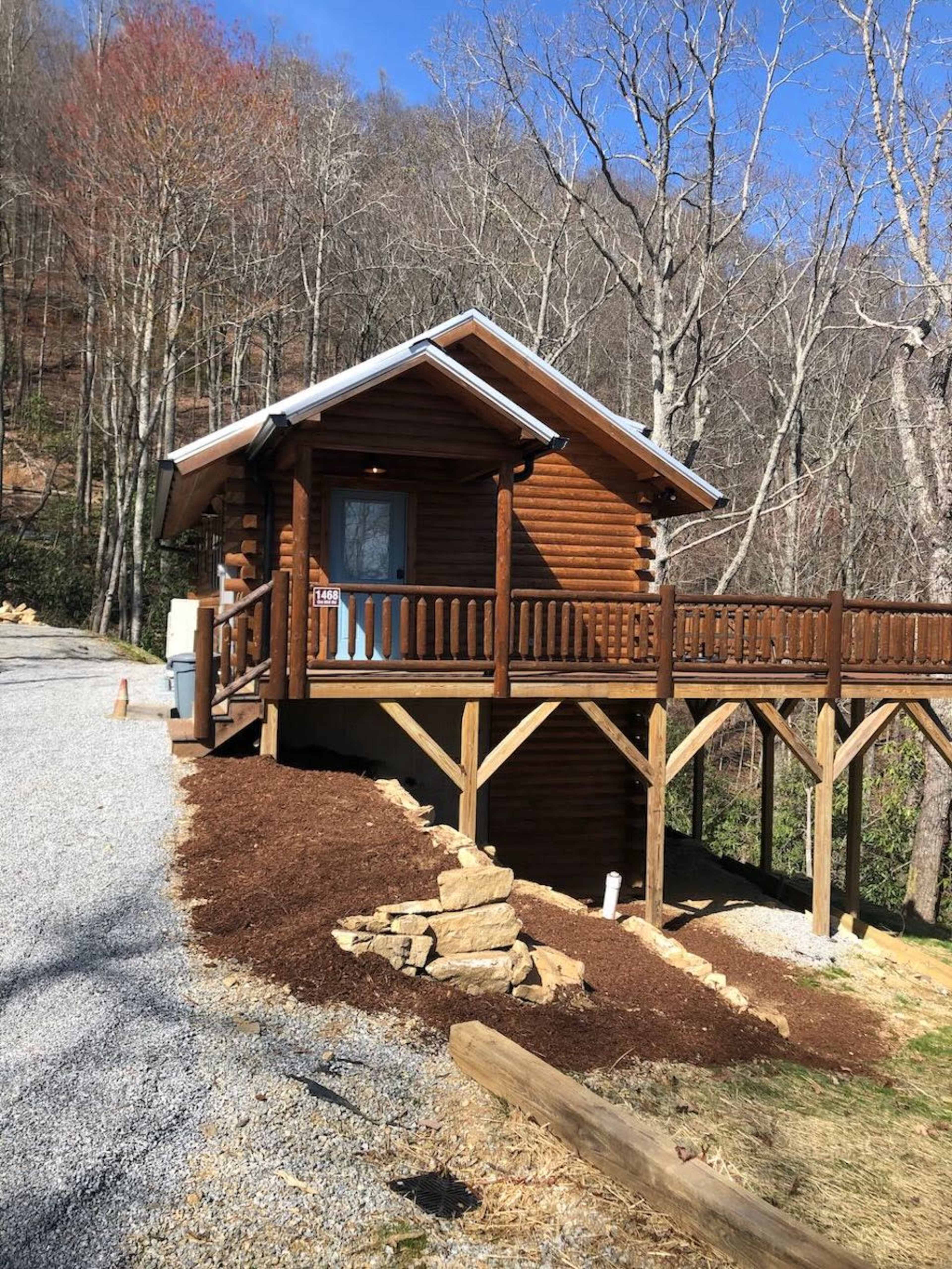 A wooden cabin with a wraparound porch is situated on a gravel driveway surrounded by trees.