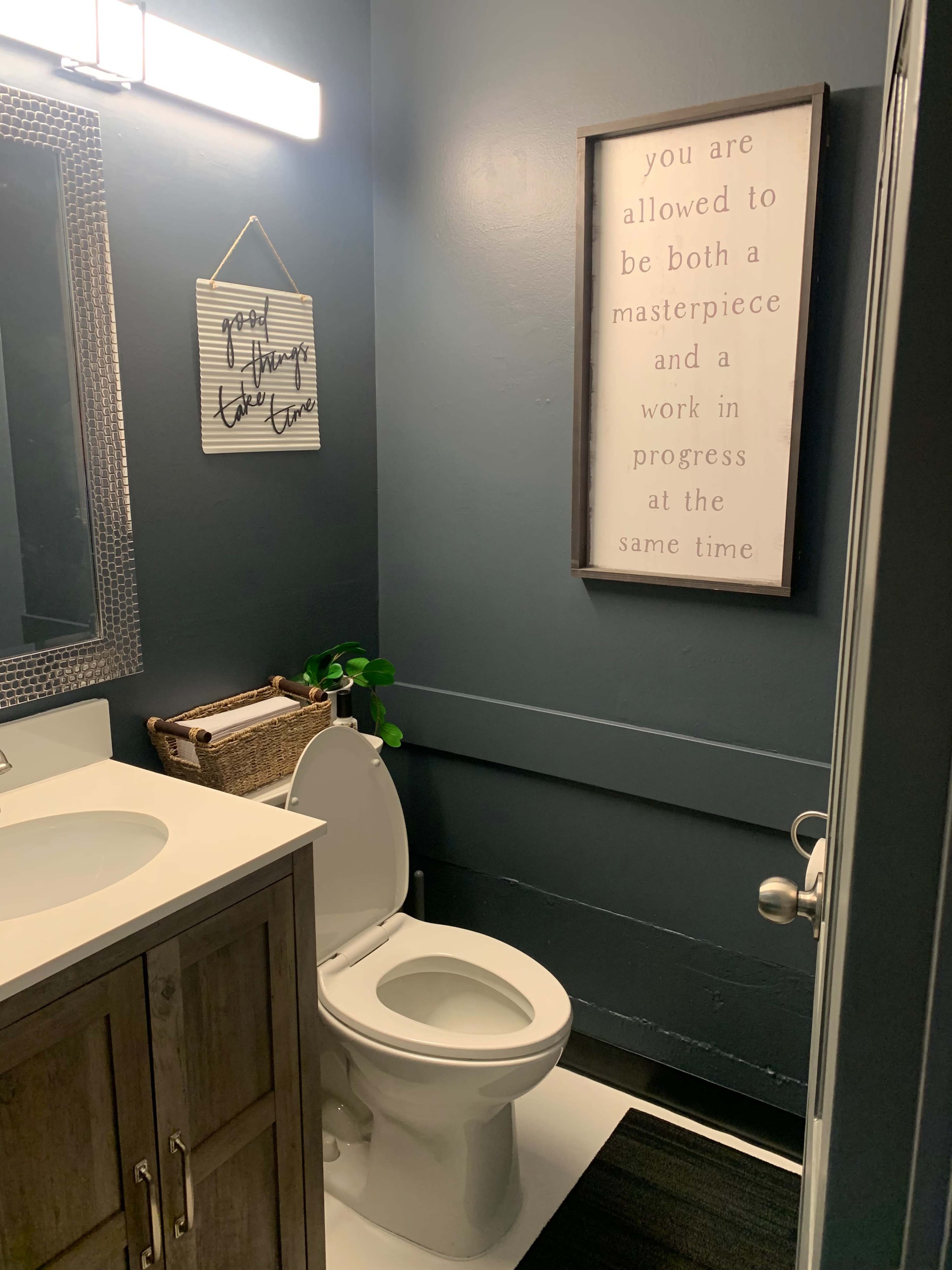 A small bathroom features a dark blue wall, a white sink, a toilet, and framed wall art, along with a decorative plant and a woven basket.