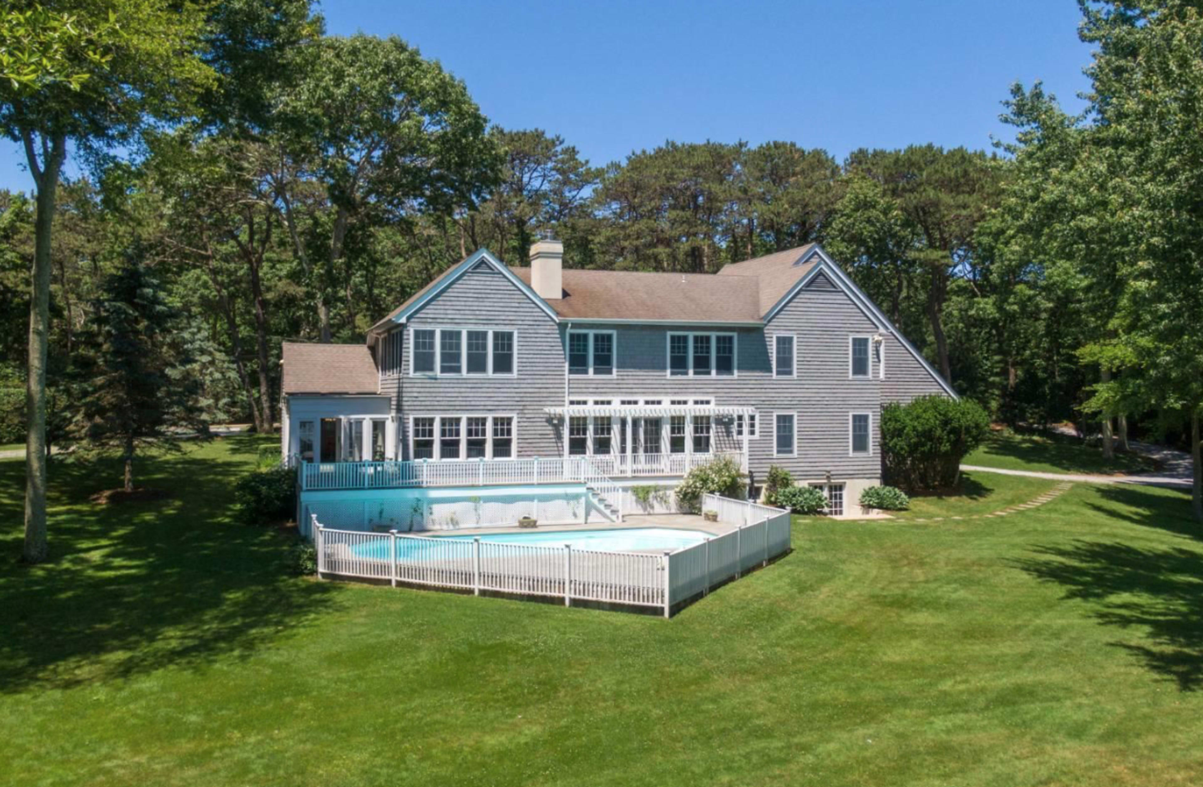 A large gray house with multiple windows and a swimming pool surrounded by a white fence sits on a grassy lawn, bordered by trees.