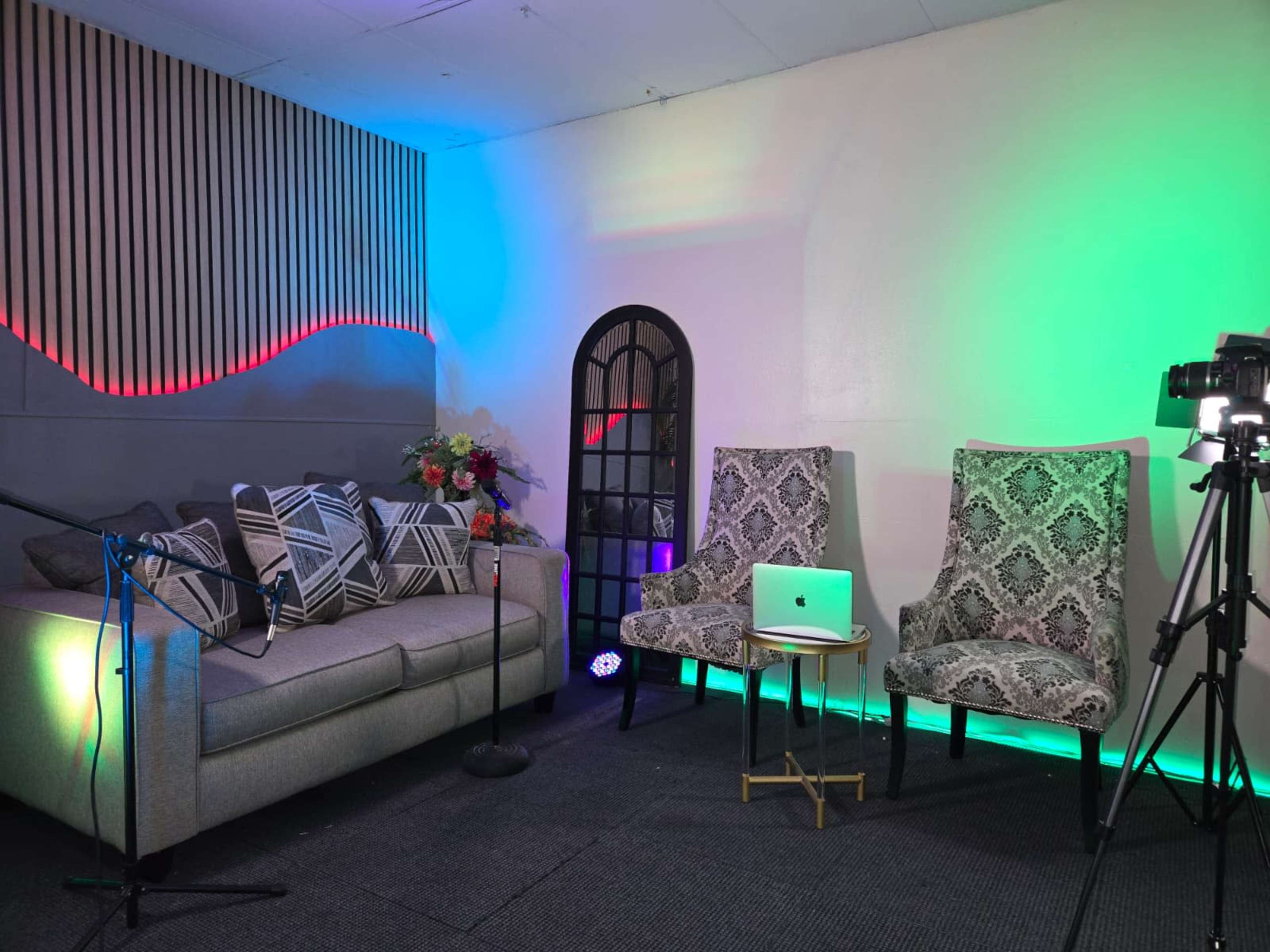 The image shows a modern studio setup with a gray sofa, two patterned chairs, a small table with a laptop, and colorful mood lighting.