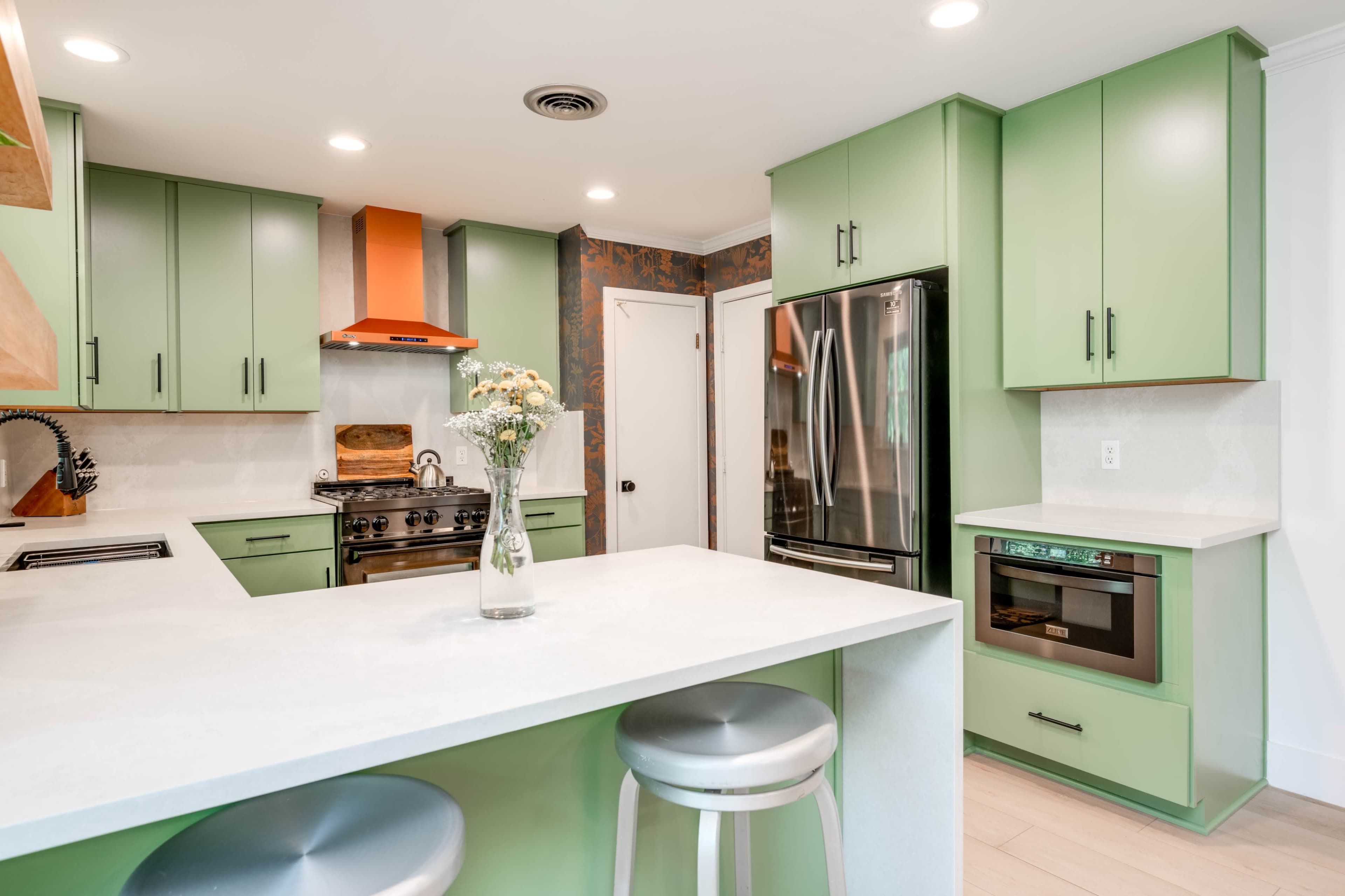 A modern kitchen features green cabinets, a stainless steel refrigerator, an orange hood above the stove, and a central island with seating.
