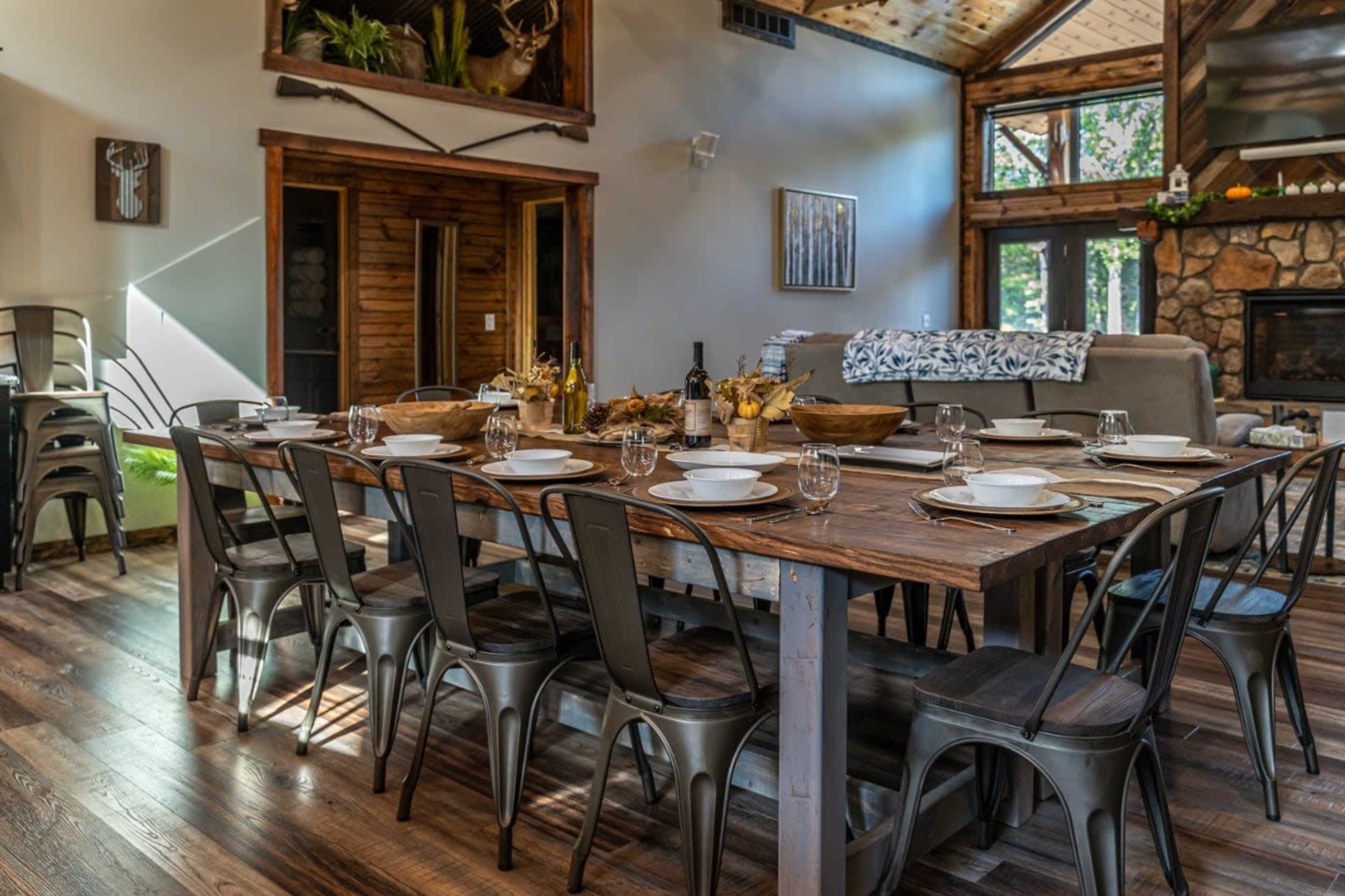 A long wooden dining table is set with plates and glassware in a rustic, well-lit room featuring large windows and a stone fireplace.