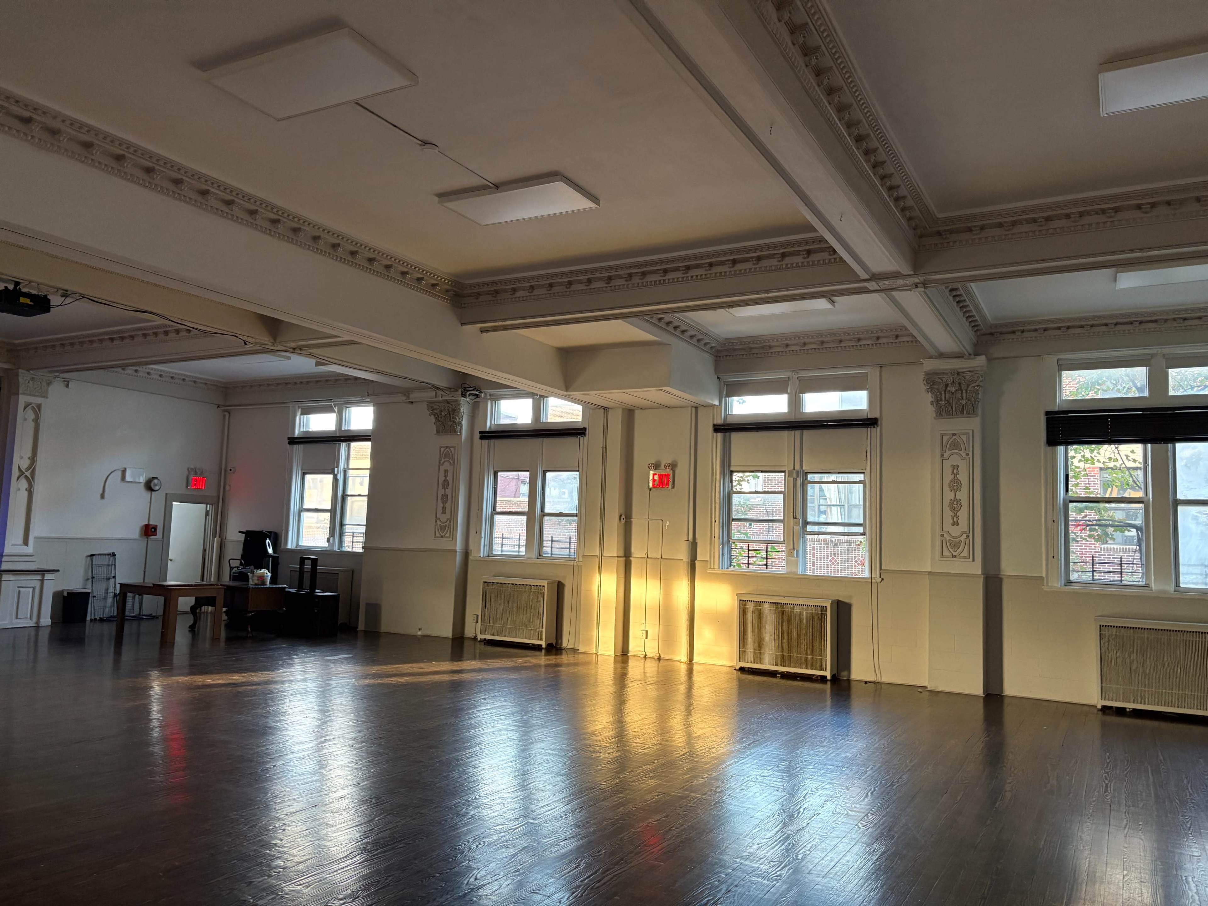 The image shows a spacious, well-lit room with wooden flooring, large windows, and a few scattered furnishings, including a table and chairs.