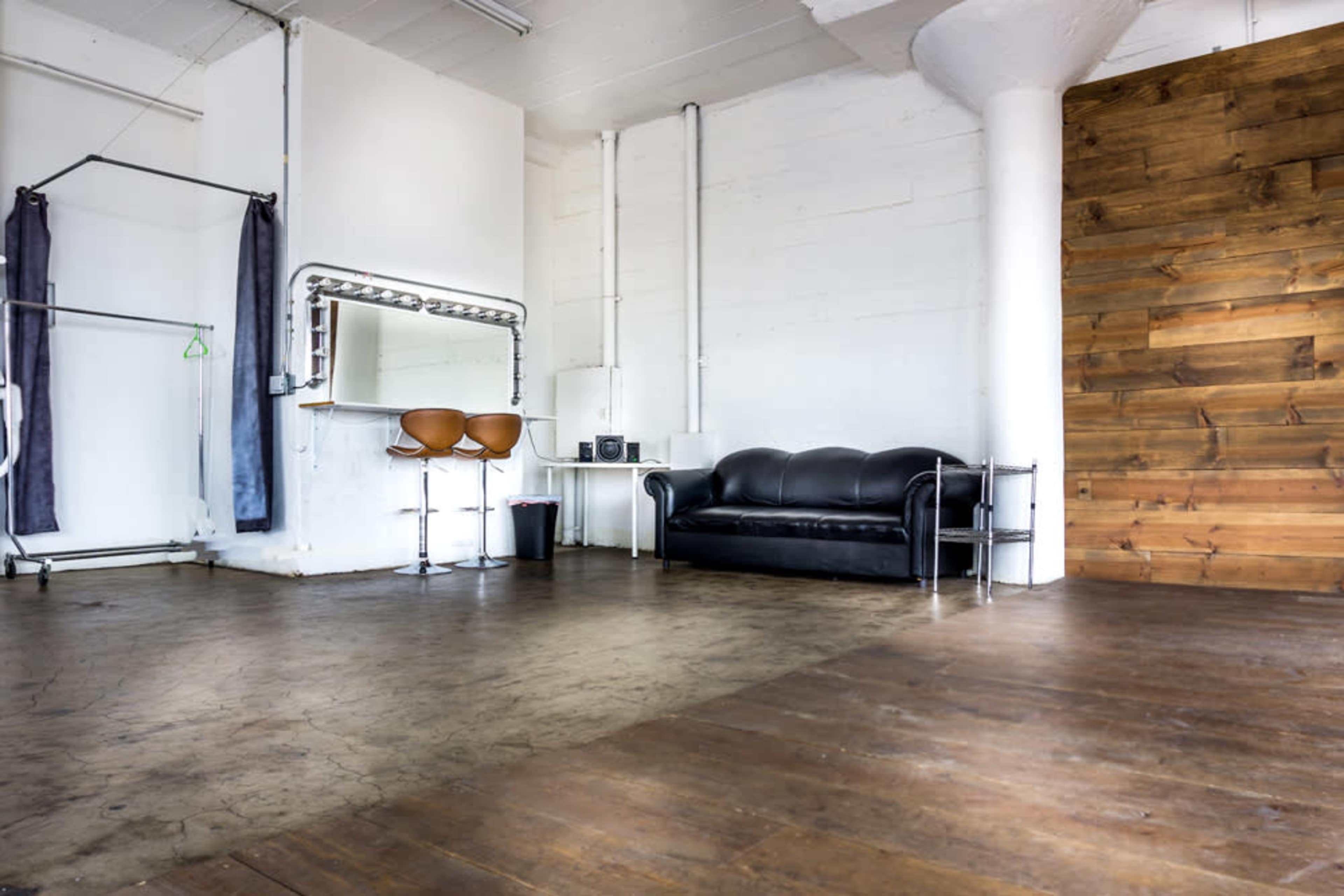The image shows a spacious interior with a black couch, a mirror surrounded by lights, two bar stools, and wooden paneling on one wall.