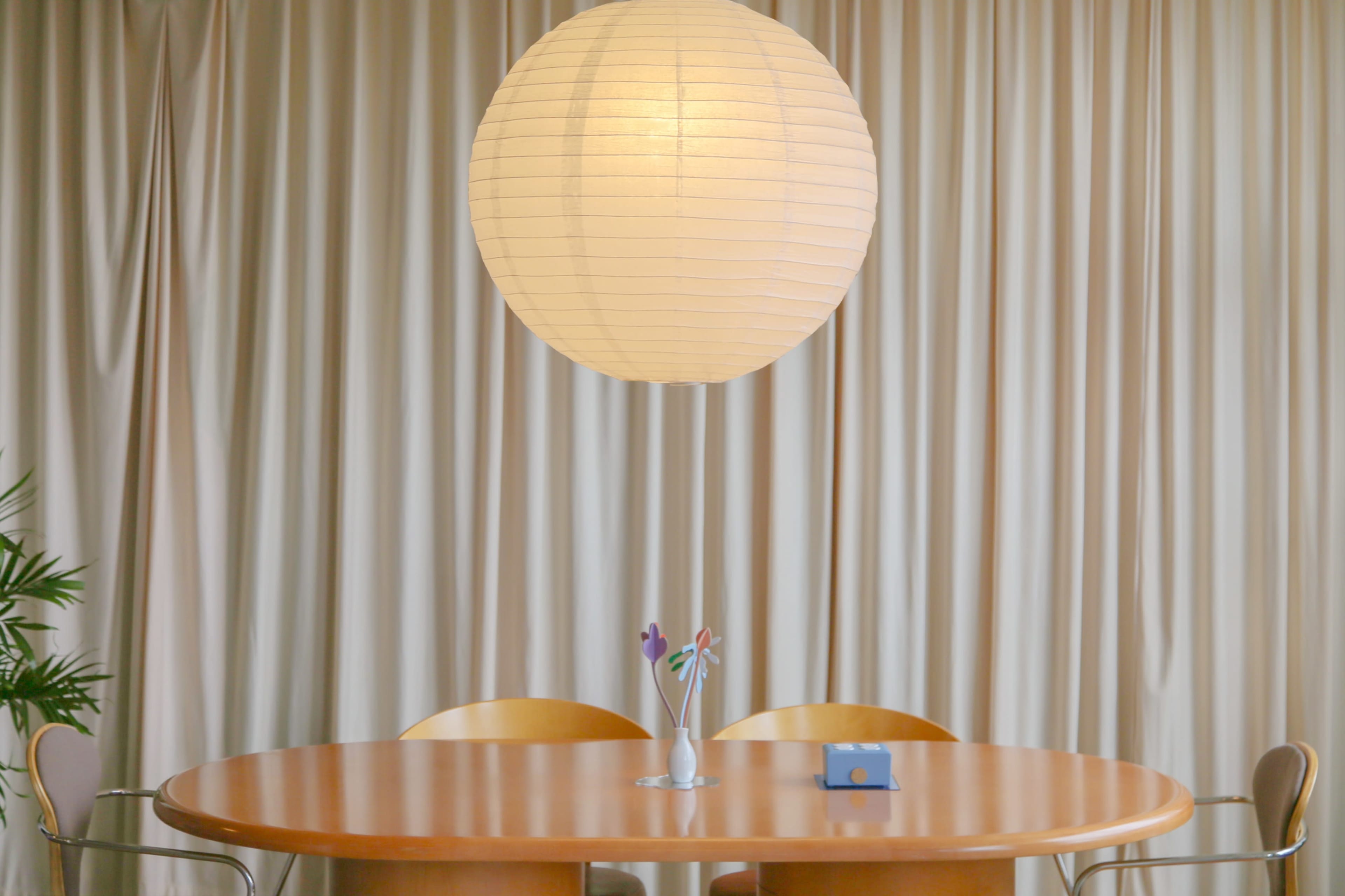 A round wooden dining table with two chairs is set beneath a large paper lantern, surrounded by beige drapes.