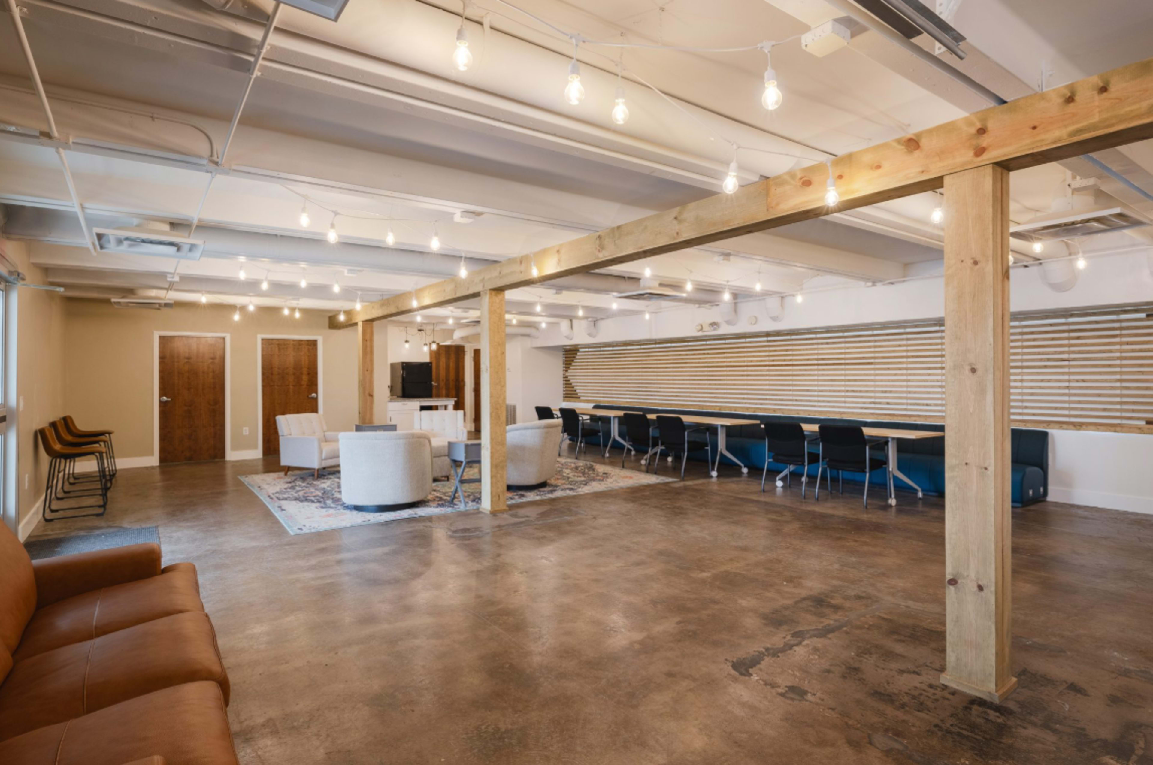 The image shows an open, modern workspace featuring brown leather seating, wooden beams, a large table with chairs, and dim pendant lighting.