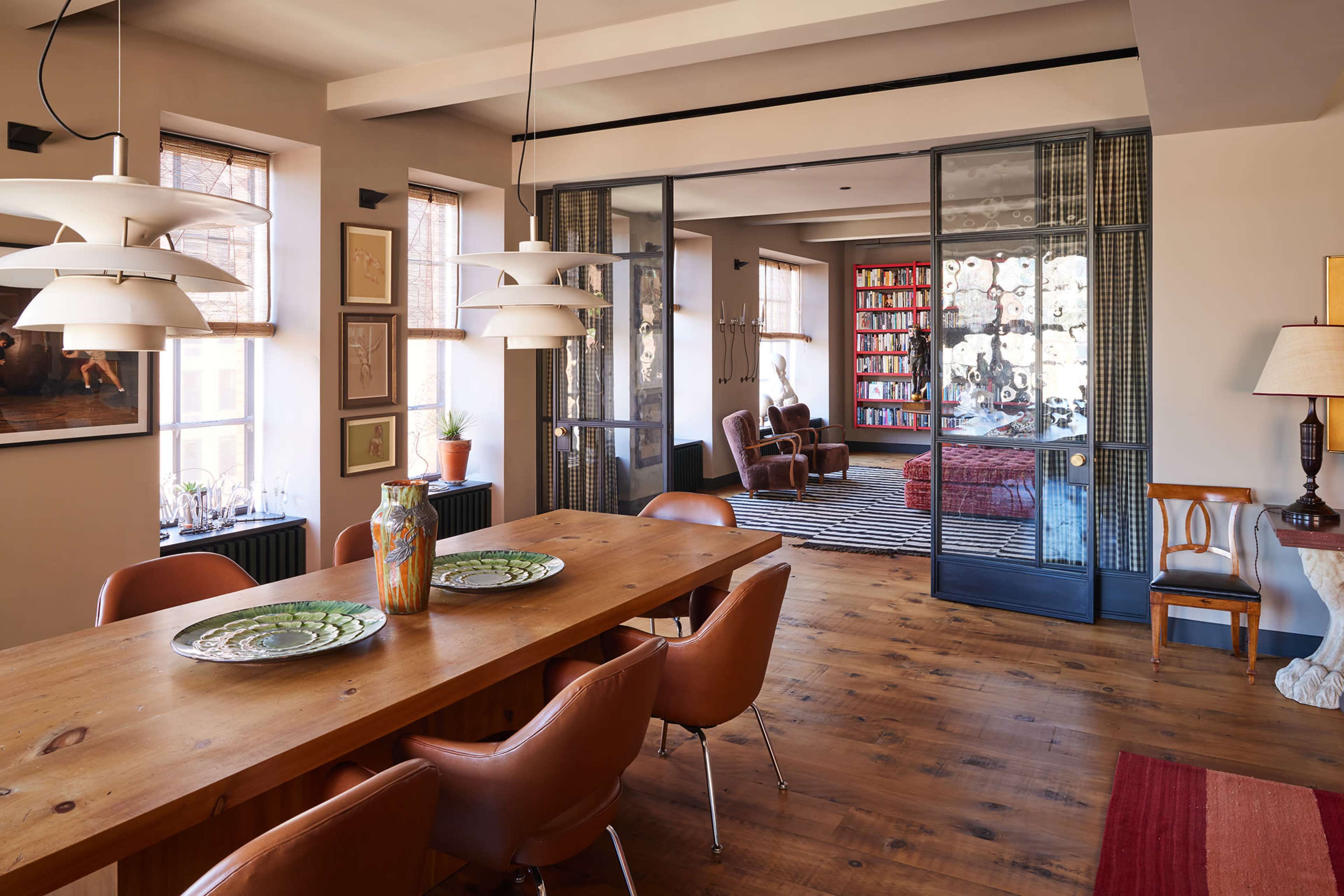 The image shows a dining area with a long wooden table and leather chairs, leading into a living space filled with bookshelves and comfortable seating.