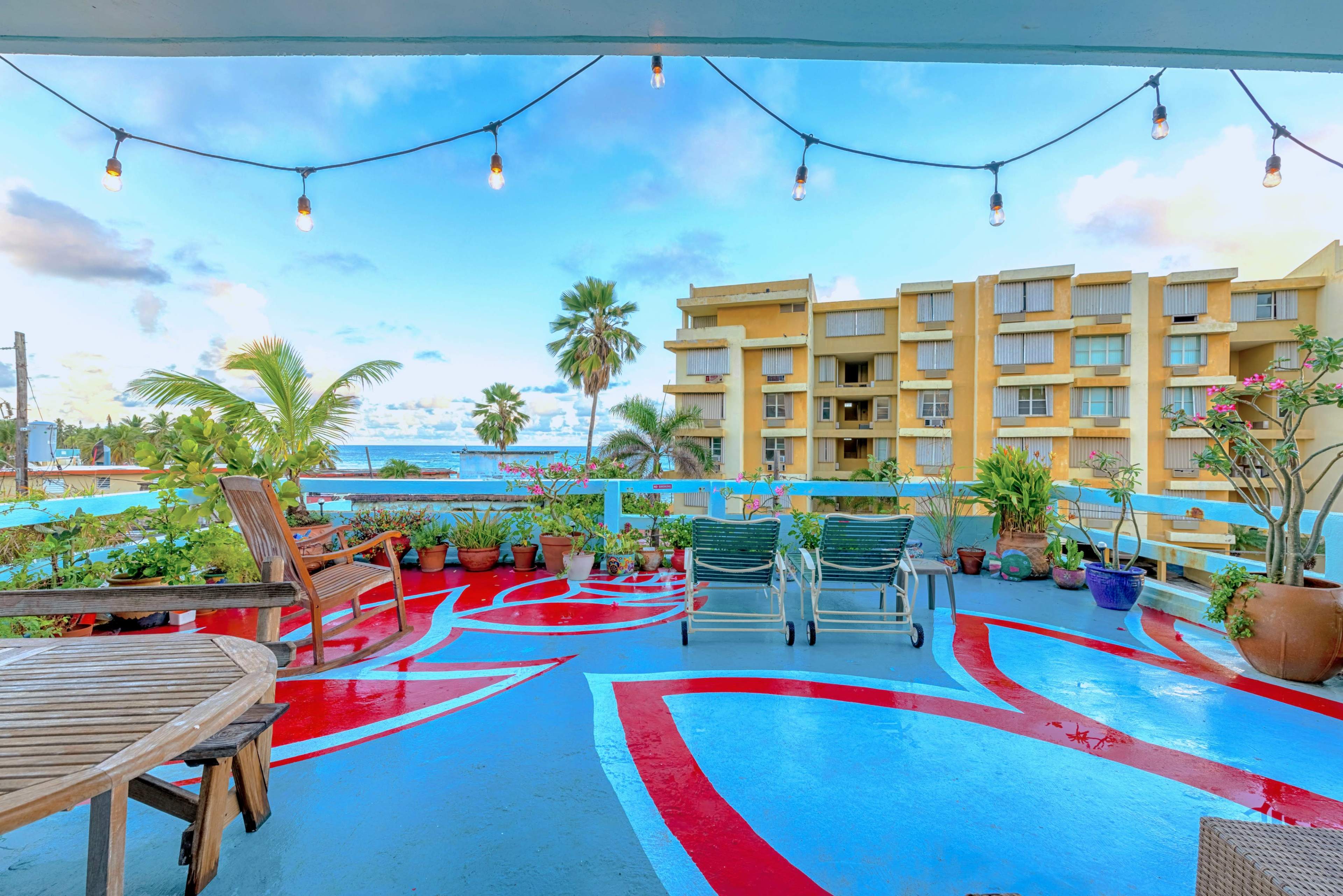 A balcony features potted plants and string lights, overlooking a body of water and a multi-story building.