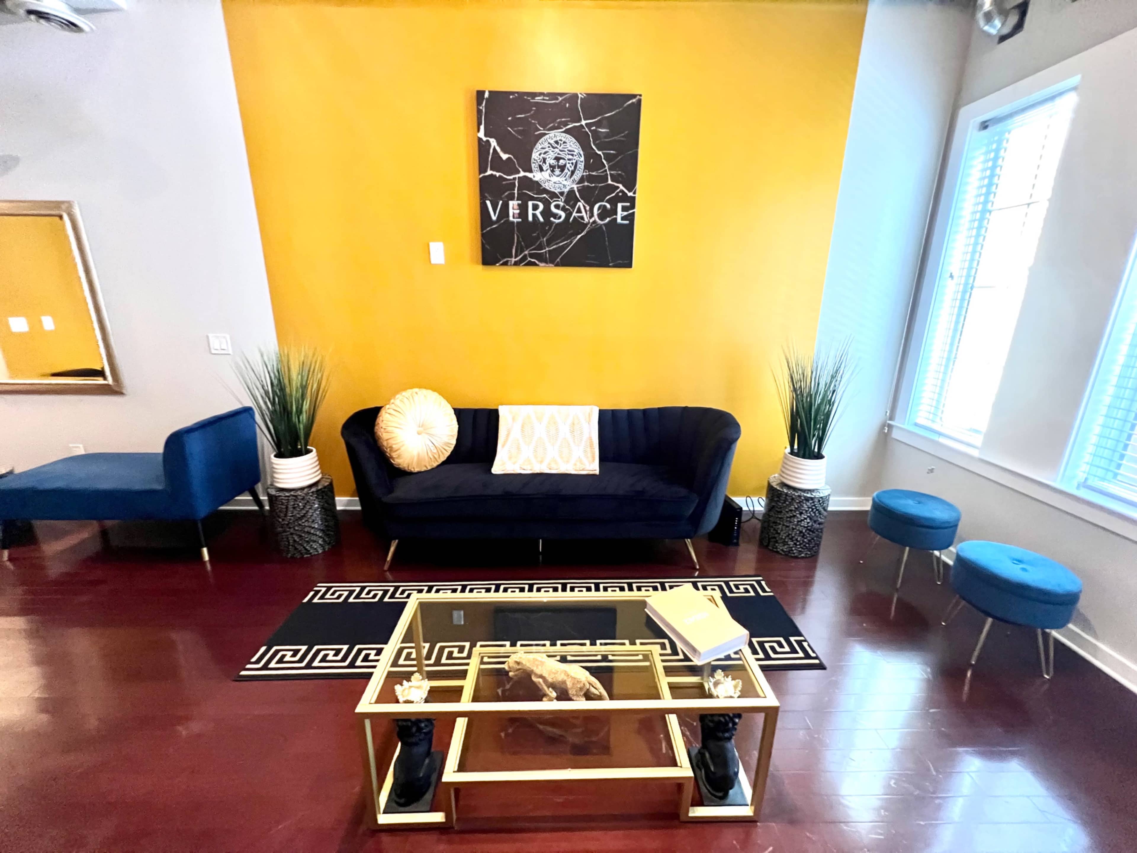 A living room features a navy blue sofa, two matching ottomans, a glass coffee table, and a large black and yellow Versace wall art above a bright yellow accent wall.