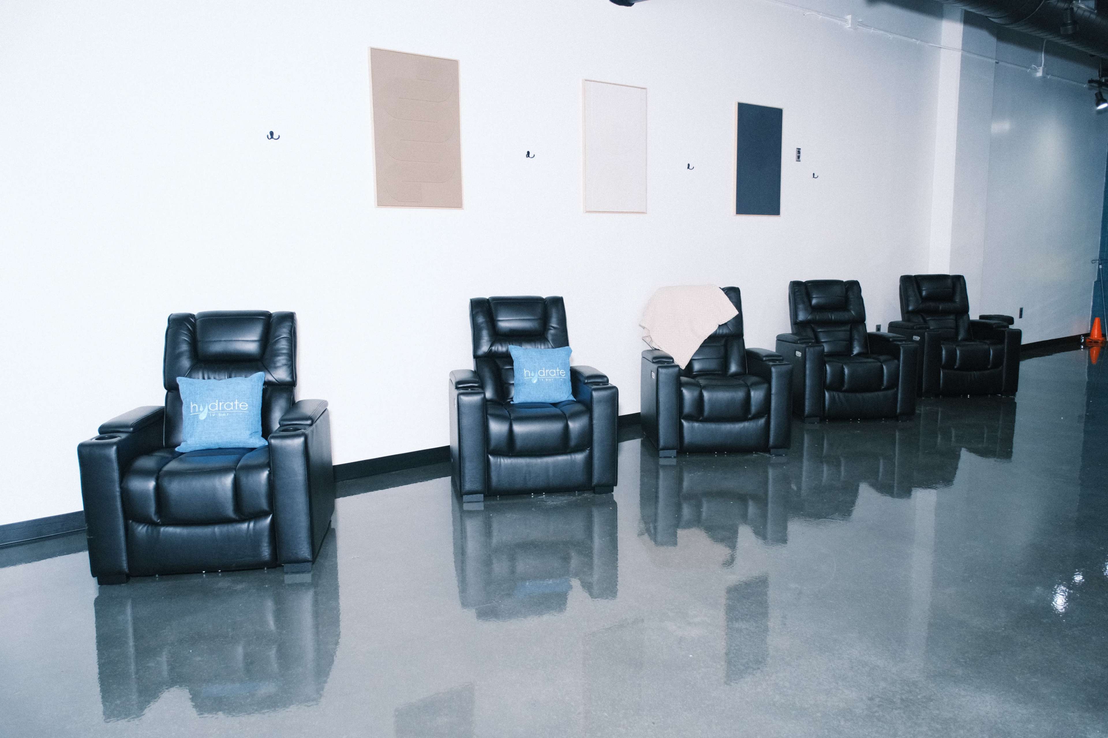 The image shows a row of five black leather recliners lined against a wall with several blank canvases hanging above them.