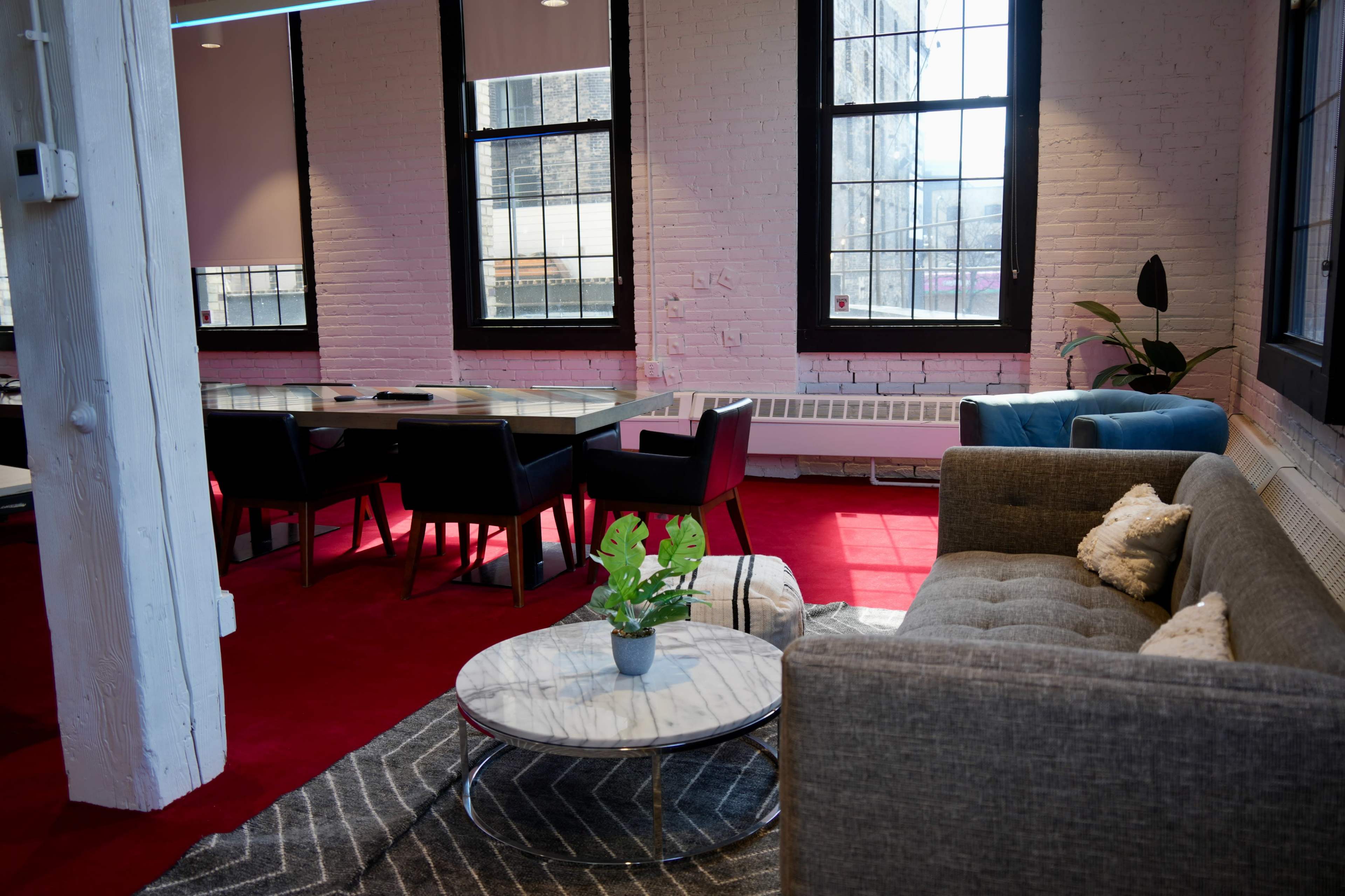The image shows a modern office space with a red carpet, a gray sofa, a marble coffee table, and a conference table surrounded by black chairs, illuminated by natural light from large windows.