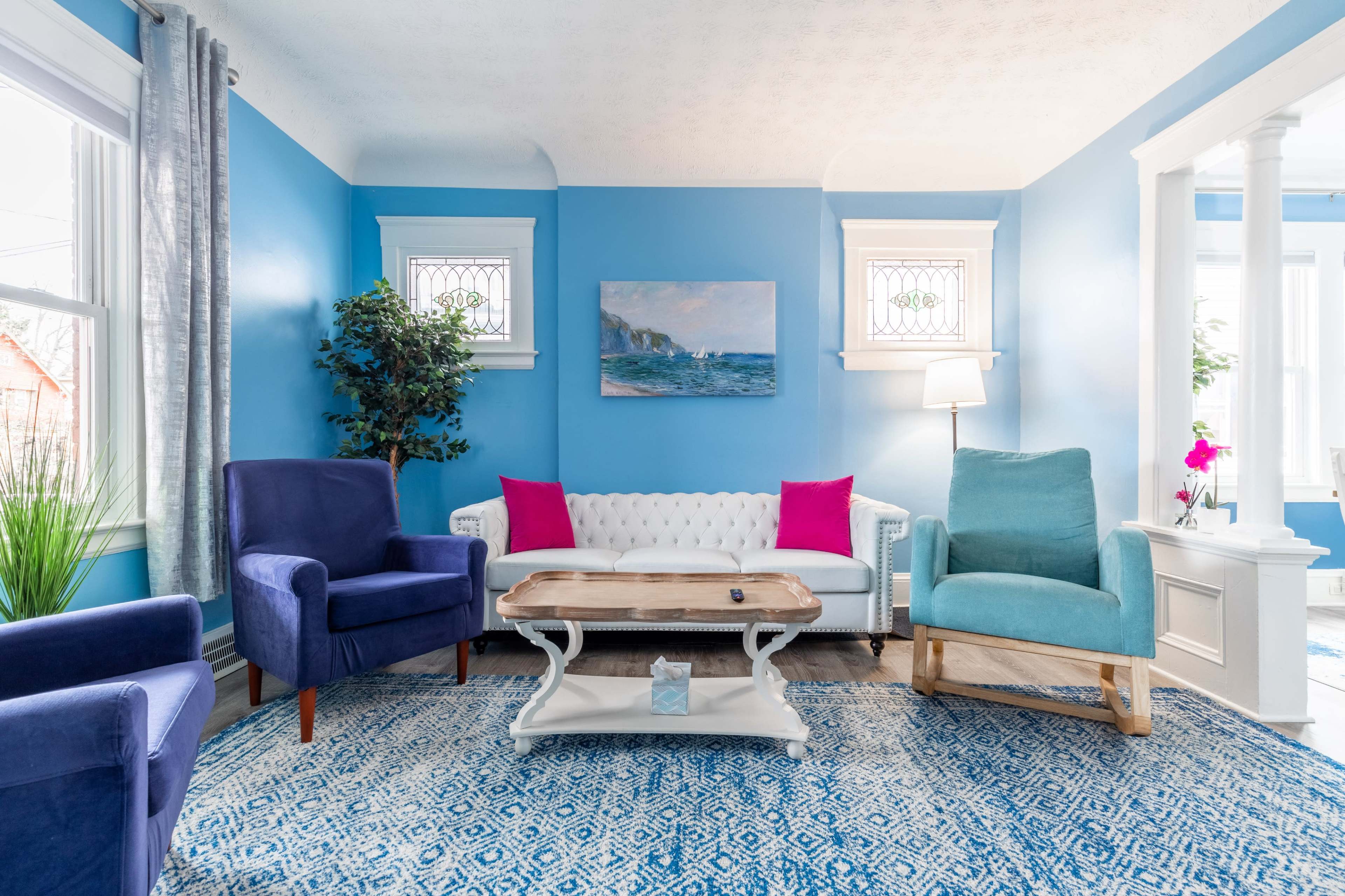 A brightly colored living room with blue walls, a white couch, and various seating options arranged around a wooden coffee table on a patterned rug.
