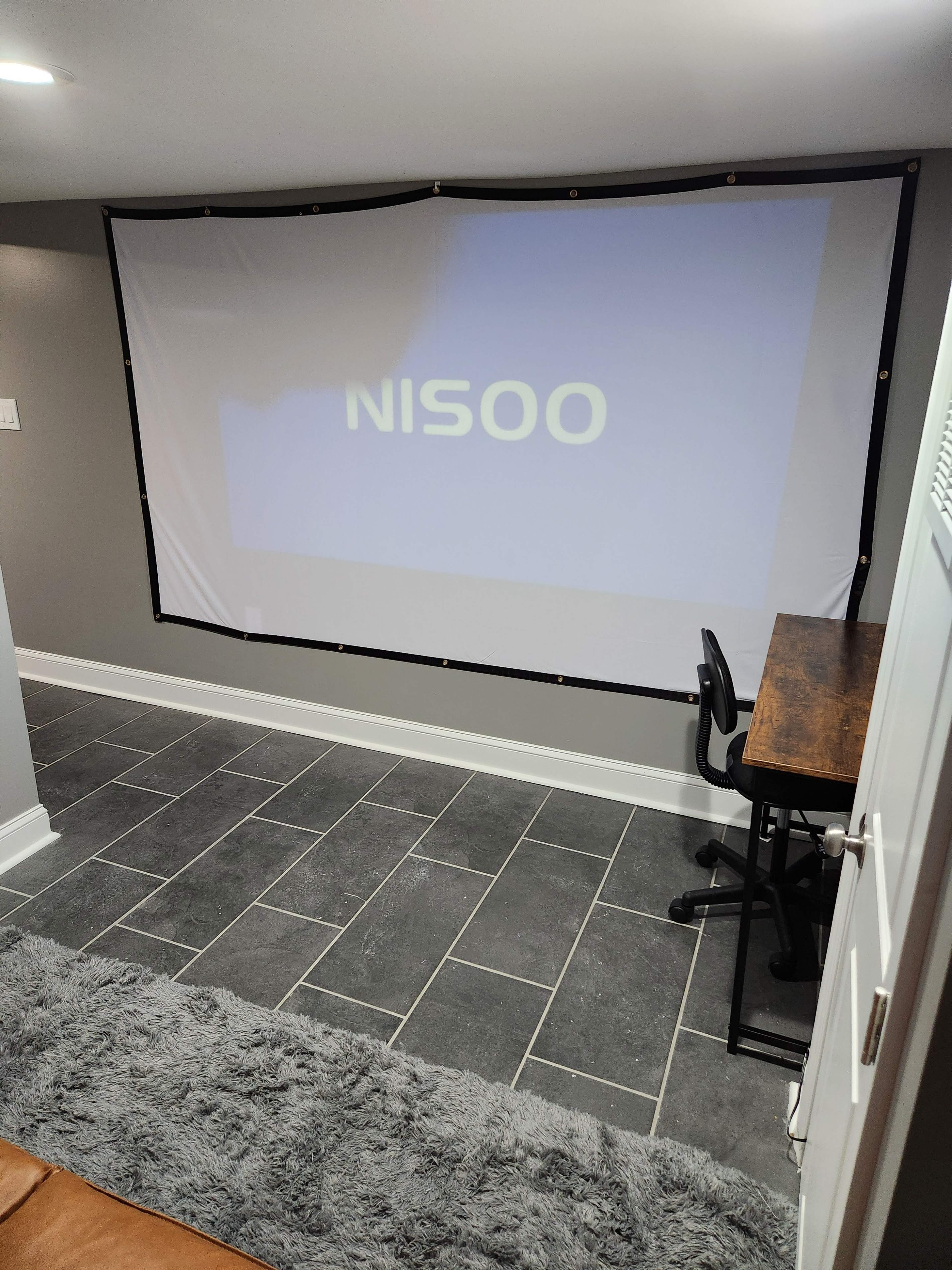 The image shows a home theater setup with a projection screen displaying the word "NISOO" on a wall, a desk with a chair, and a gray rug on the floor.
