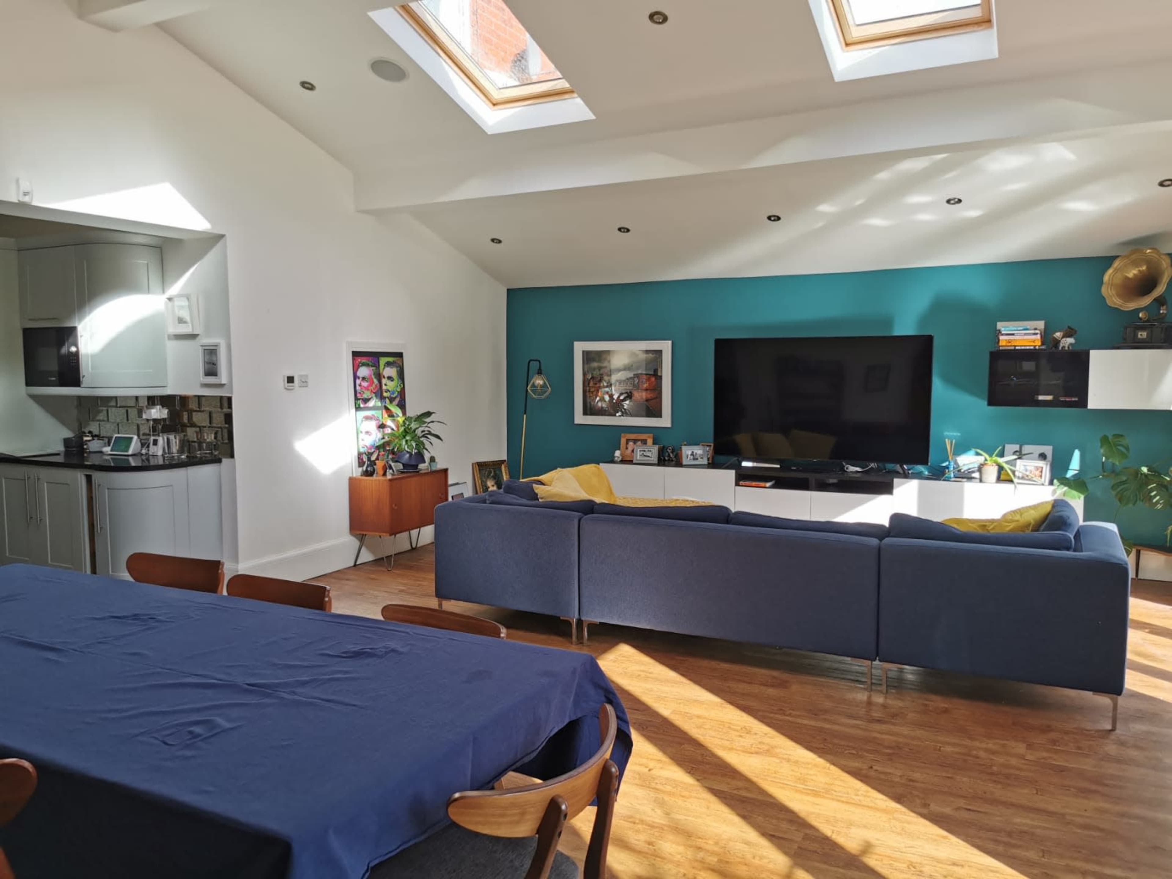 A modern living space with a blue sofa, a large television against a teal wall, a dining table, and skylights overhead.