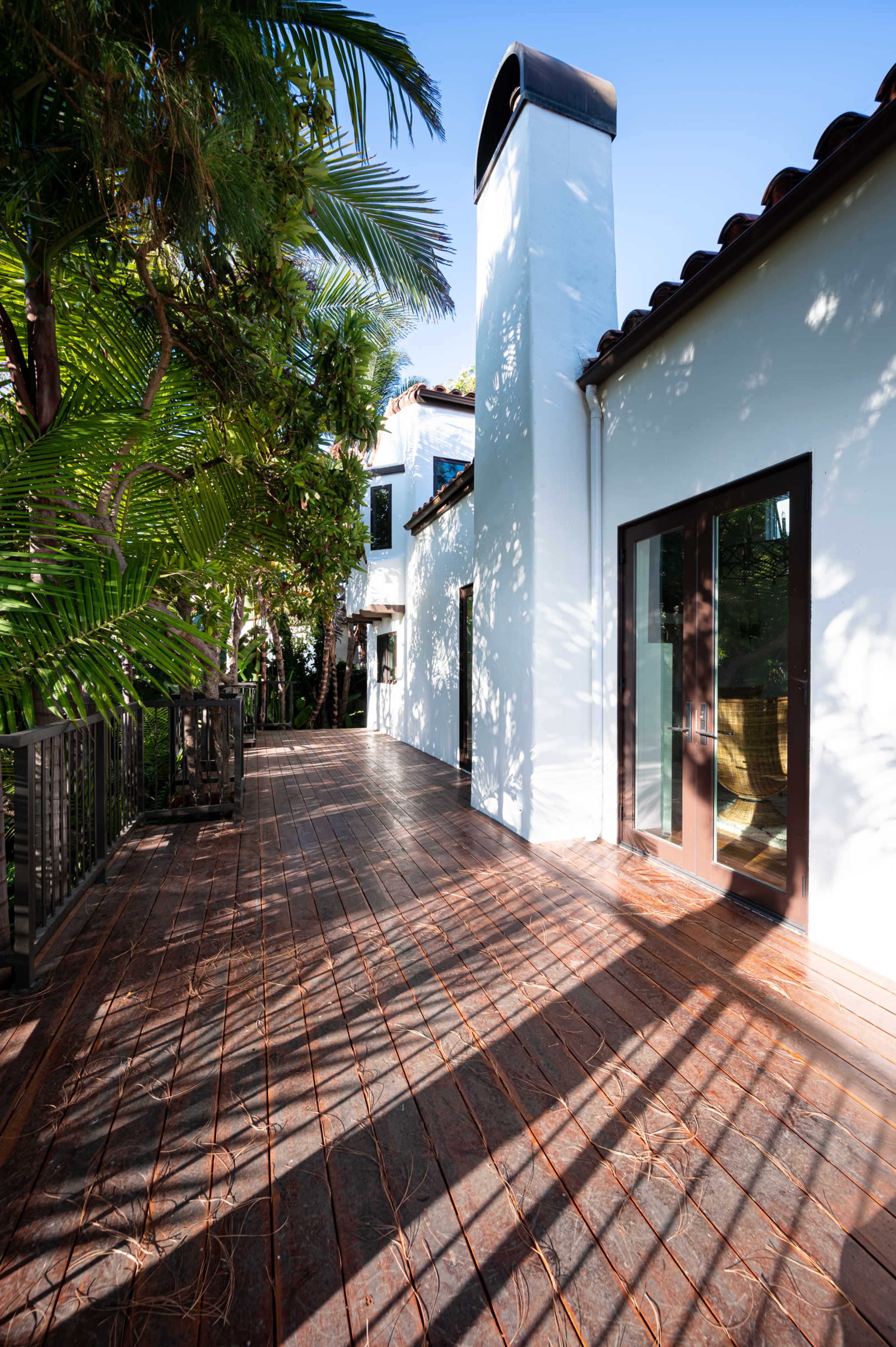A wooden deck lines the side of a white building, surrounded by lush greenery and featuring a tall chimney.