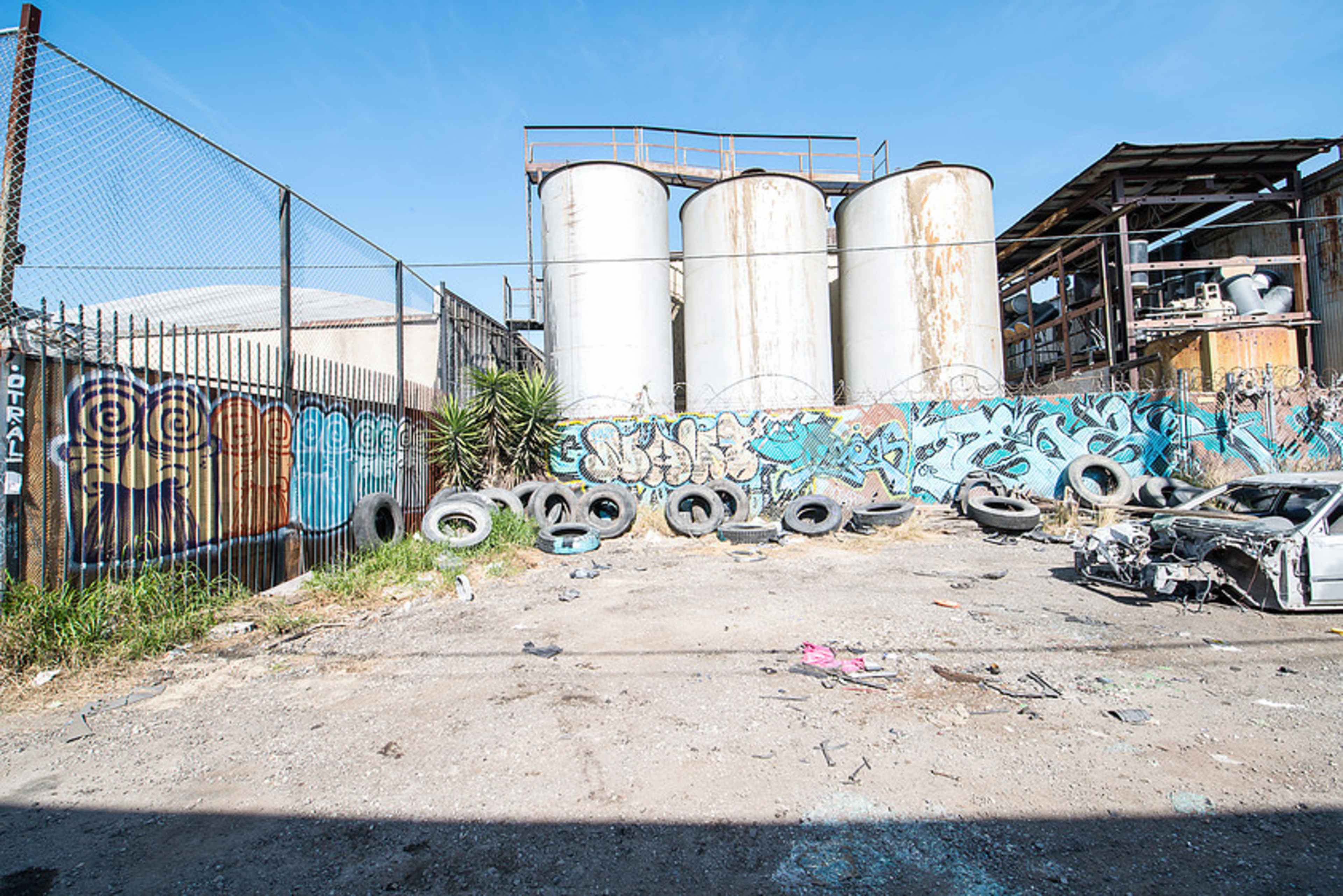 The image shows an industrial area with three large metal silos in the background, surrounded by graffiti-covered walls, discarded tires, and the remains of a car.