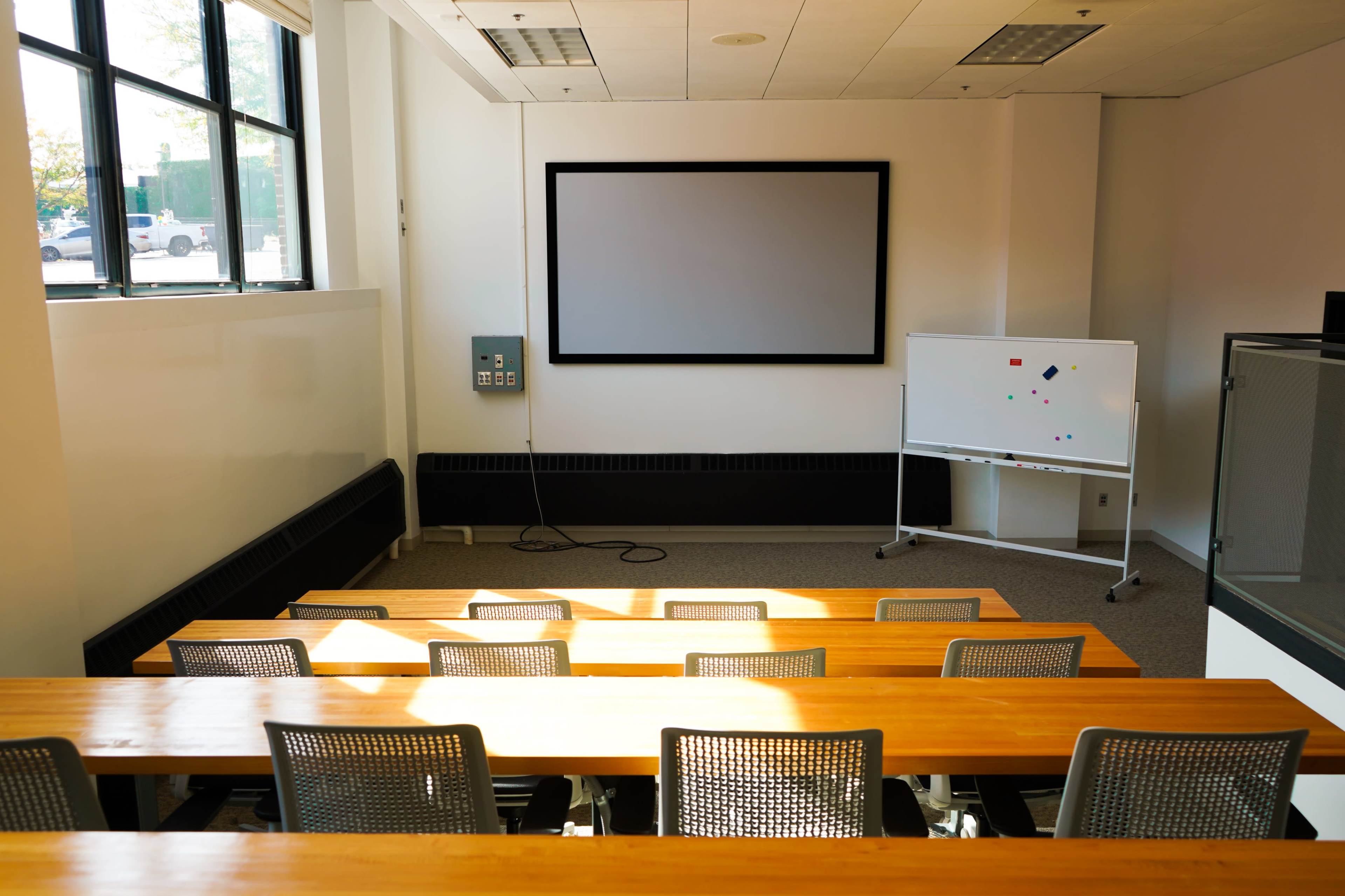 The image shows a classroom with empty wooden desks arranged in rows, a large screen on the wall, and a whiteboard at the front.