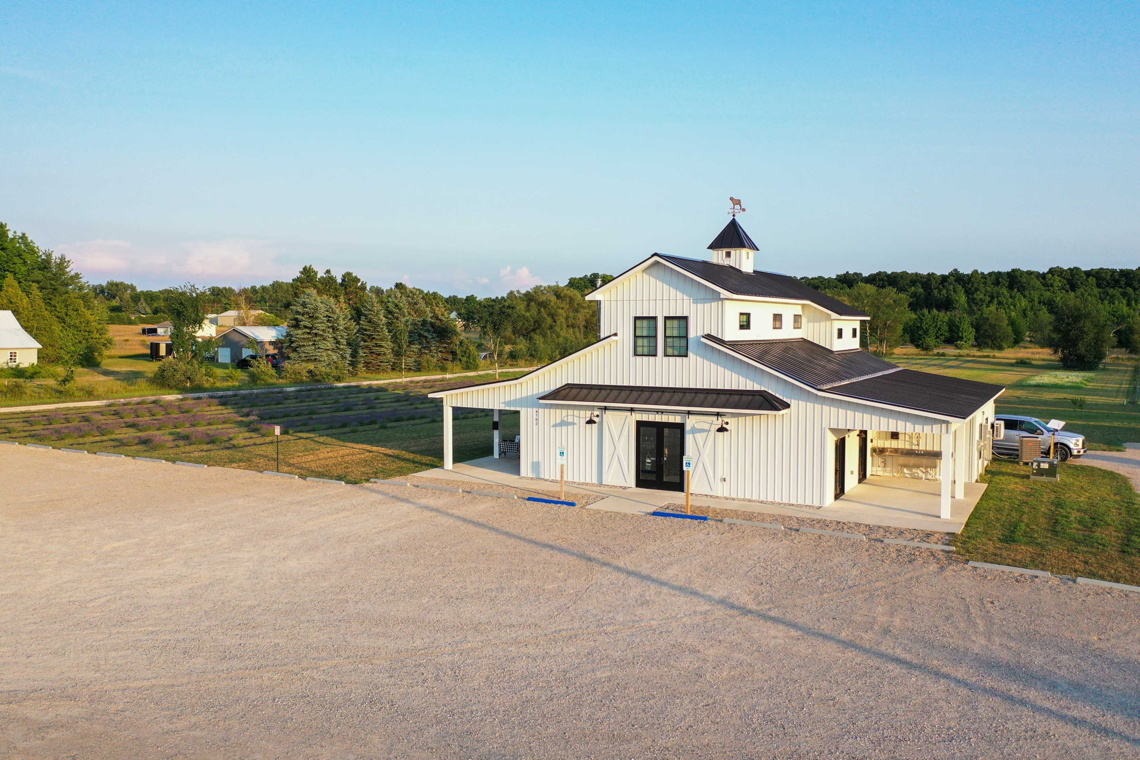 The image shows a modern white barn-style building with a black roof, surrounded by landscaped fields and trees under a clear blue sky.