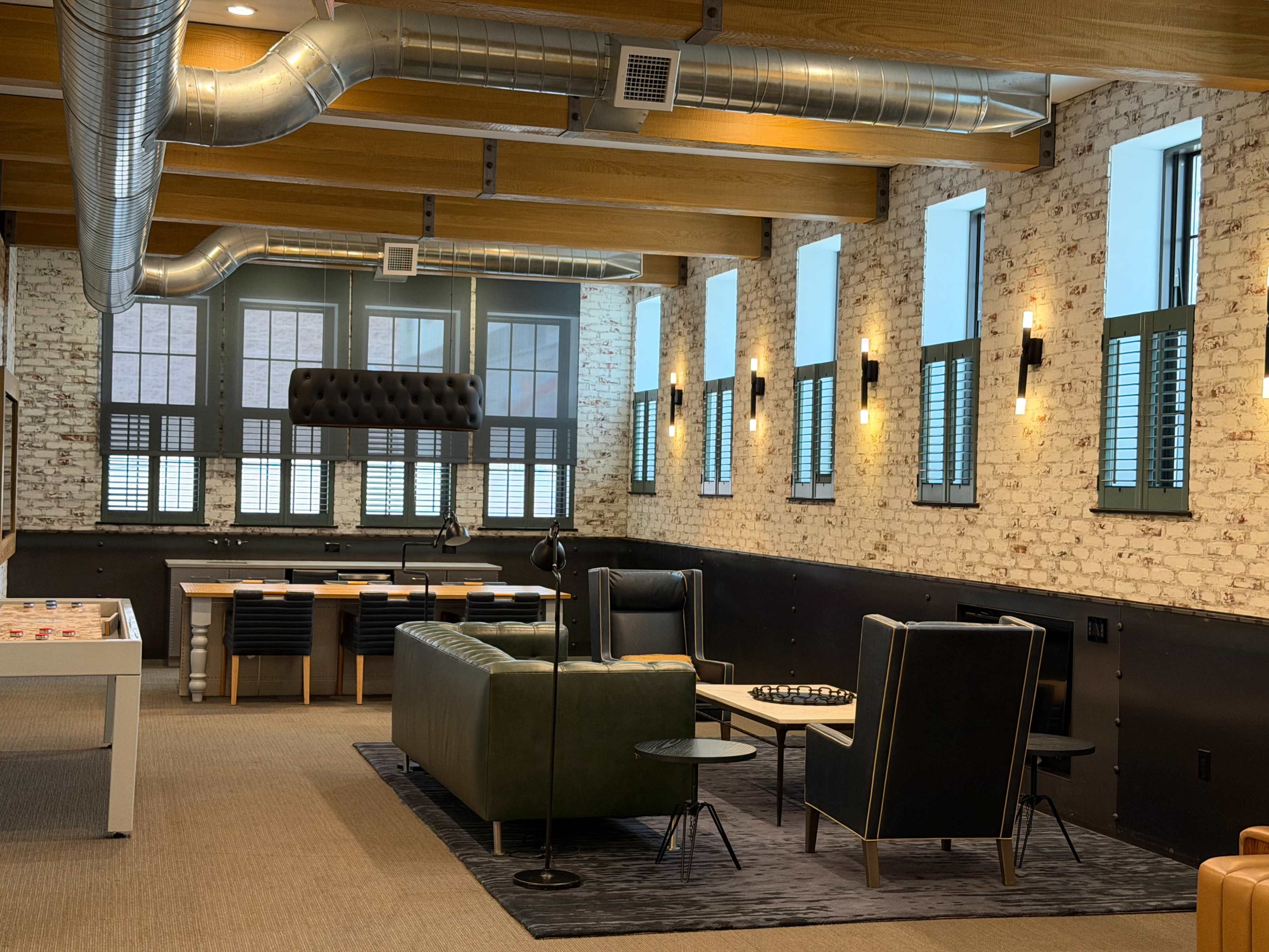 The image shows a modern lounge area with a green leather sofa, black chairs, a coffee table, and exposed brick walls, featuring large windows and metal light fixtures.