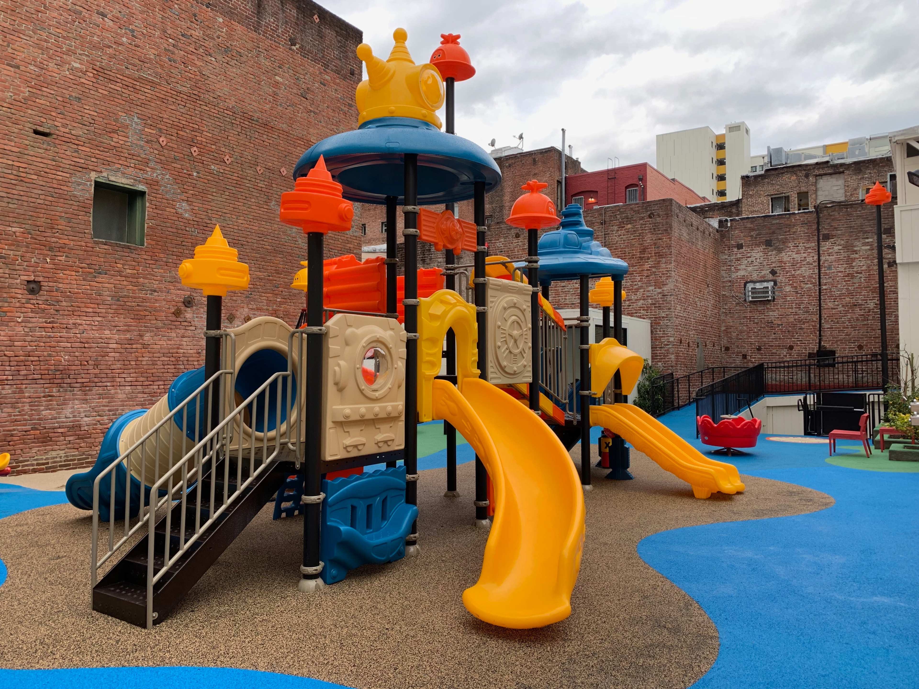 A brightly colored playground structure with slides and climbing features situated on a blue surface in an urban setting.