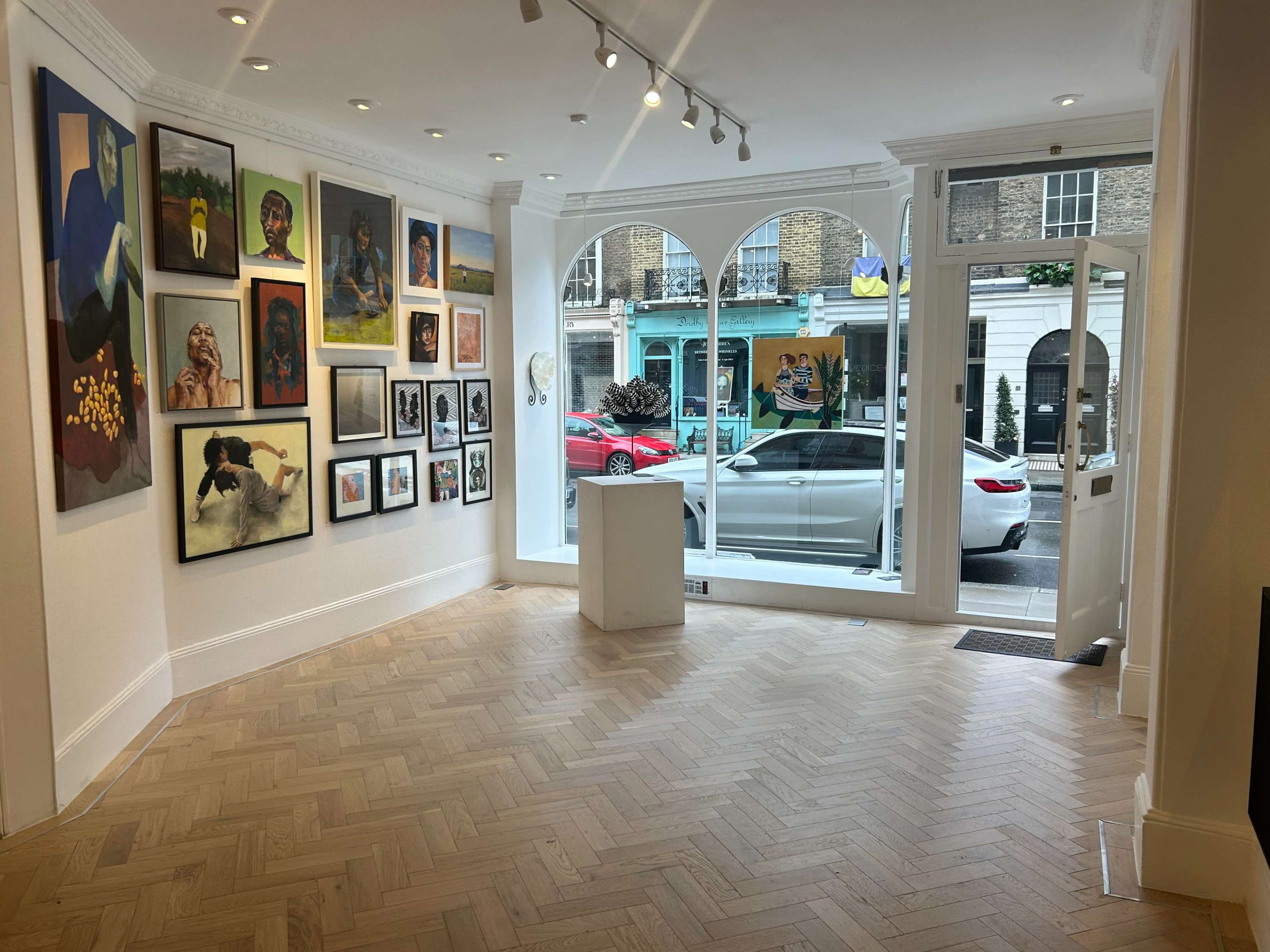 An art gallery interior featuring various framed artworks on the walls, with large windows that provide a view of the street outside.