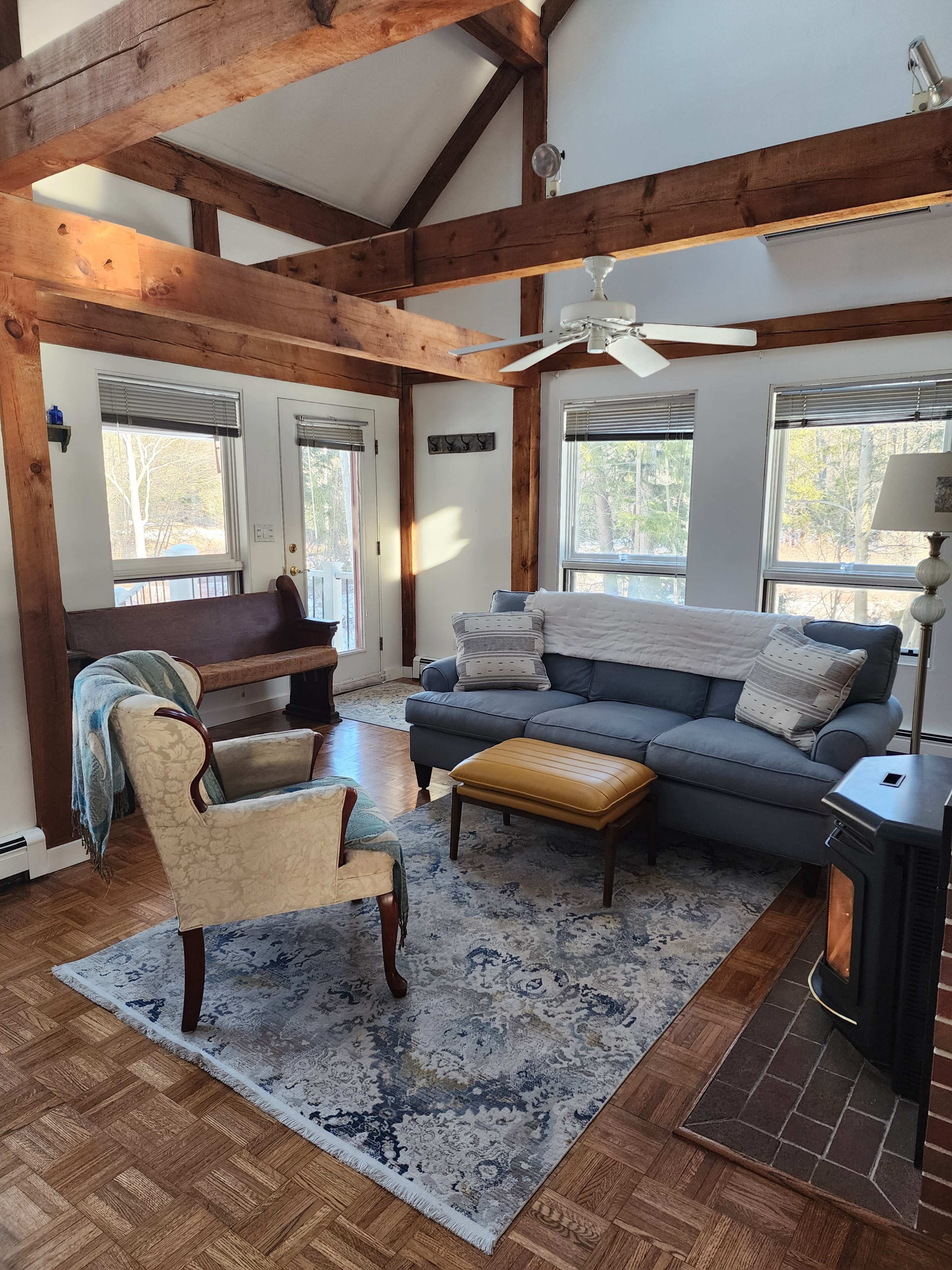 The living room features a blue sofa, a patterned area rug, and wooden beams supporting the ceiling, with a chair and a small ottoman nearby.