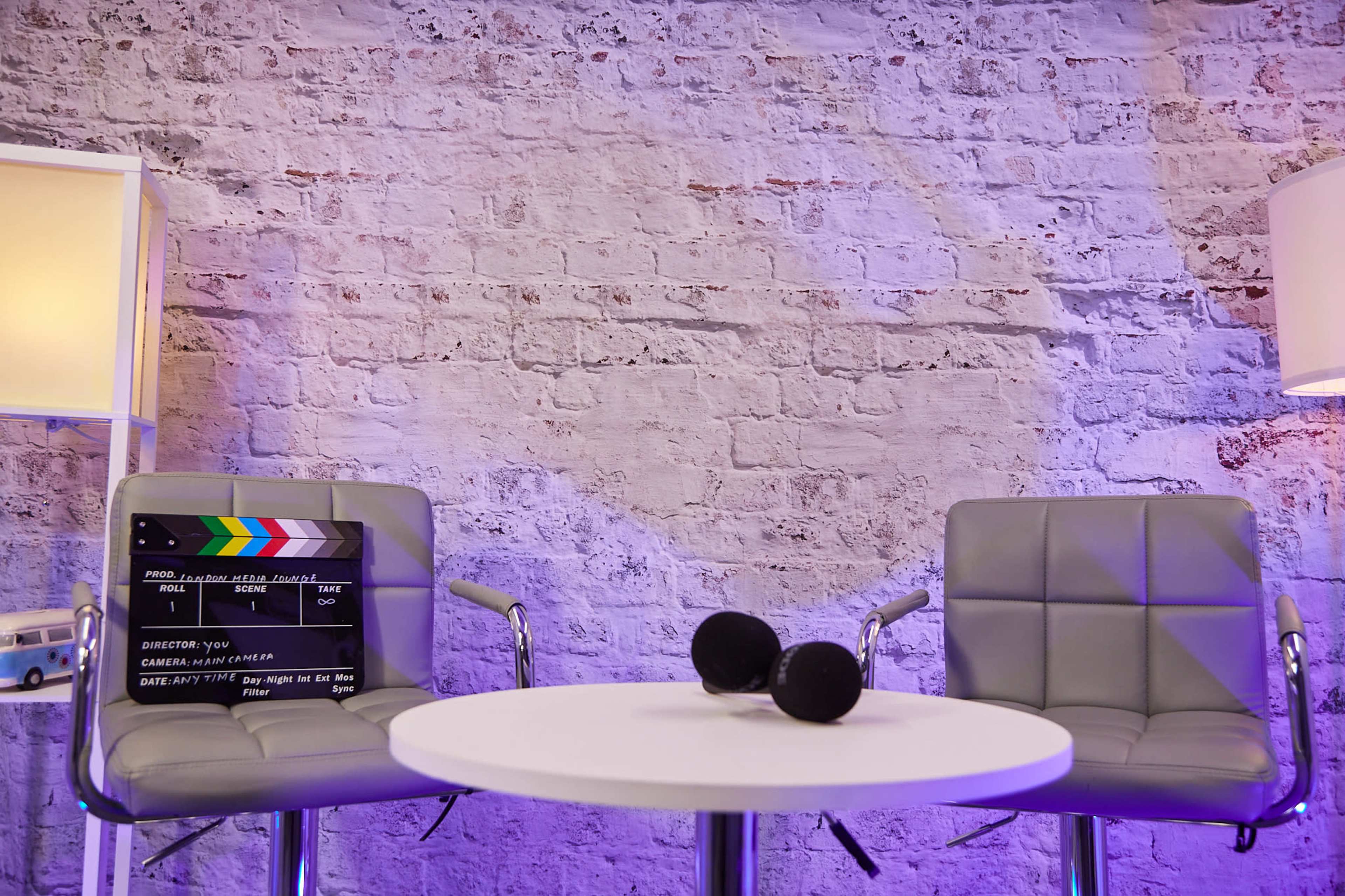 The image shows two gray chairs facing a round table with a film clapperboard and two microphones on it, set against a textured brick wall illuminated by soft lighting.