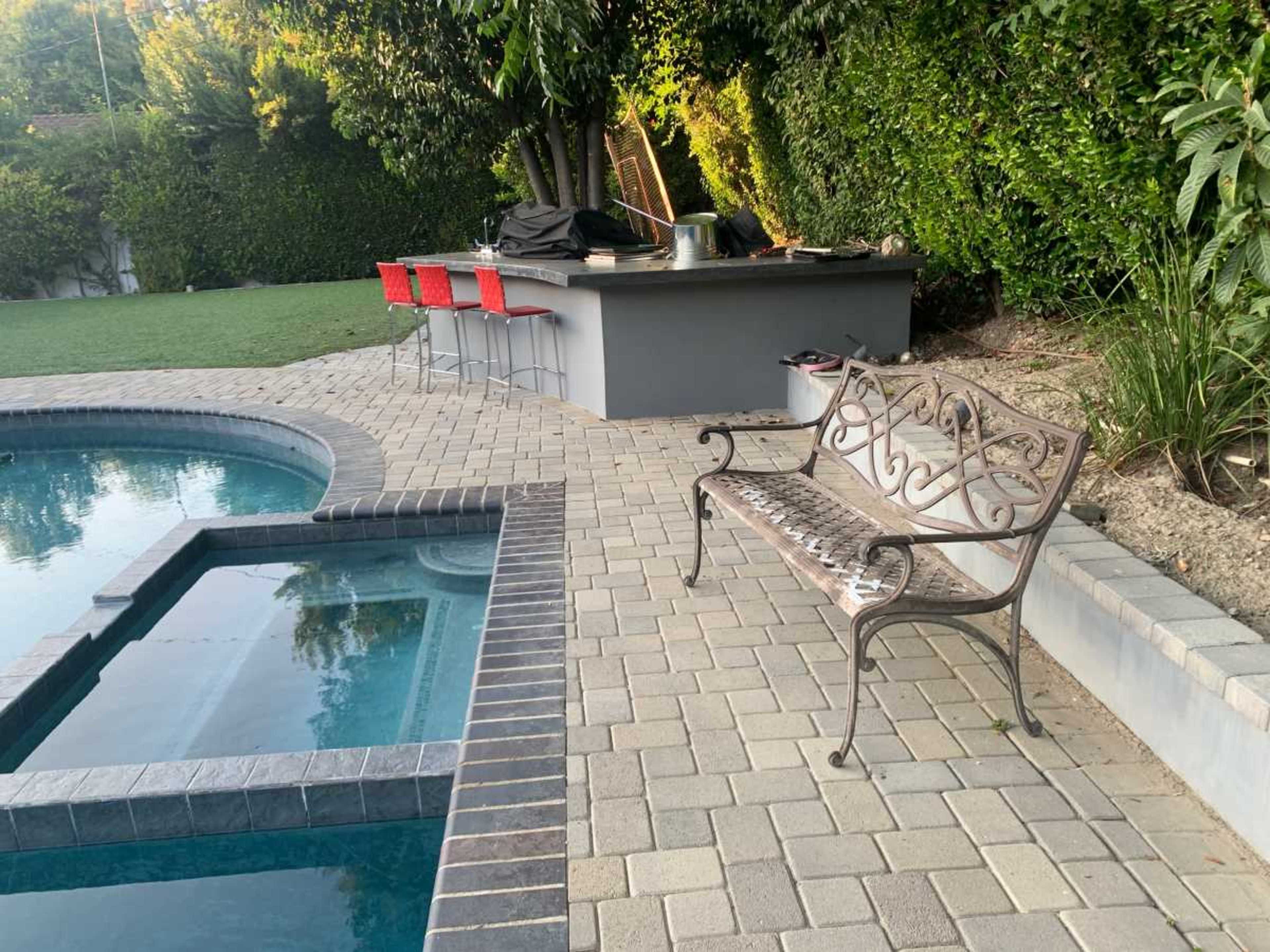 A metal bench is positioned beside a swimming pool, with a modern outdoor kitchen and red barstools visible in the background.