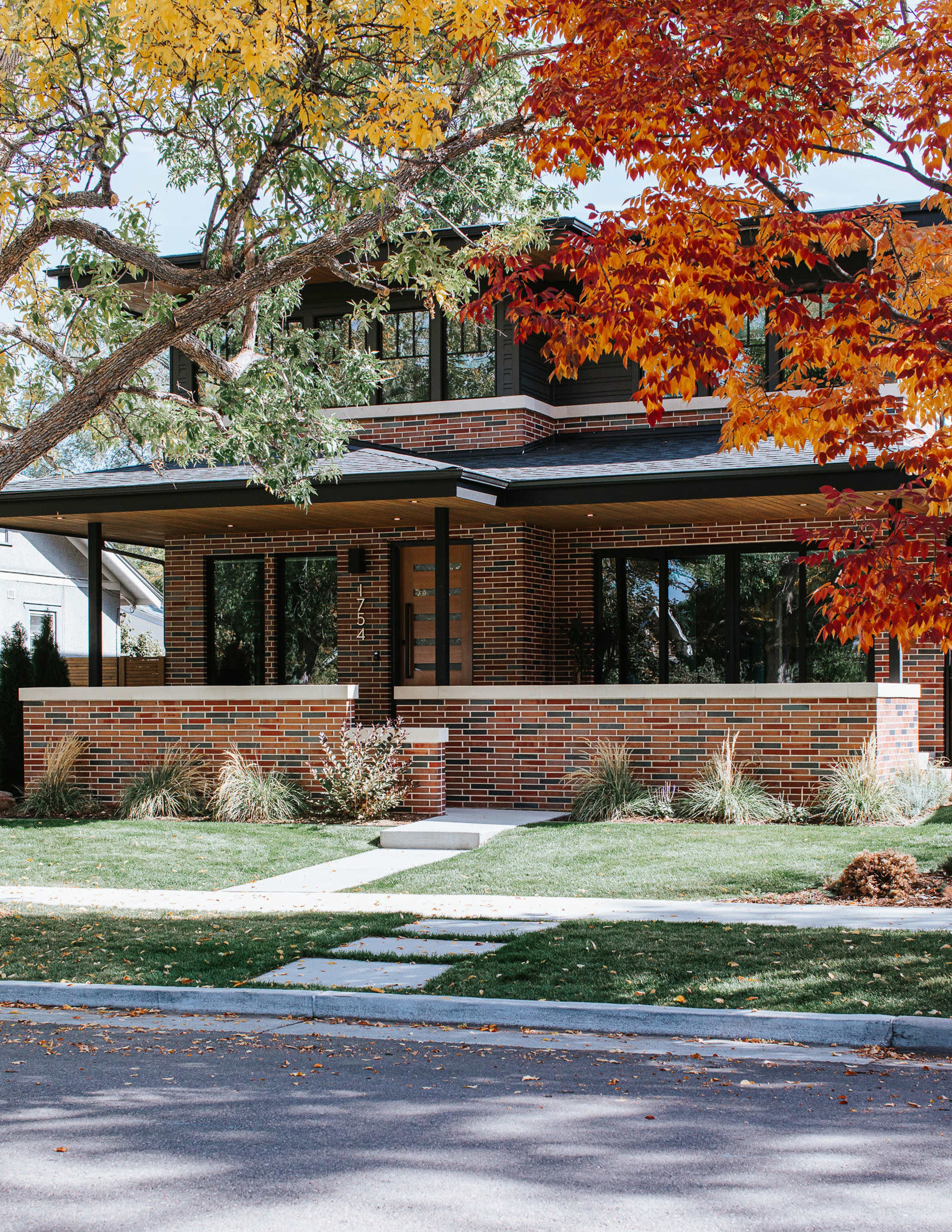 A modern brick house with large windows is framed by colorful autumn foliage.