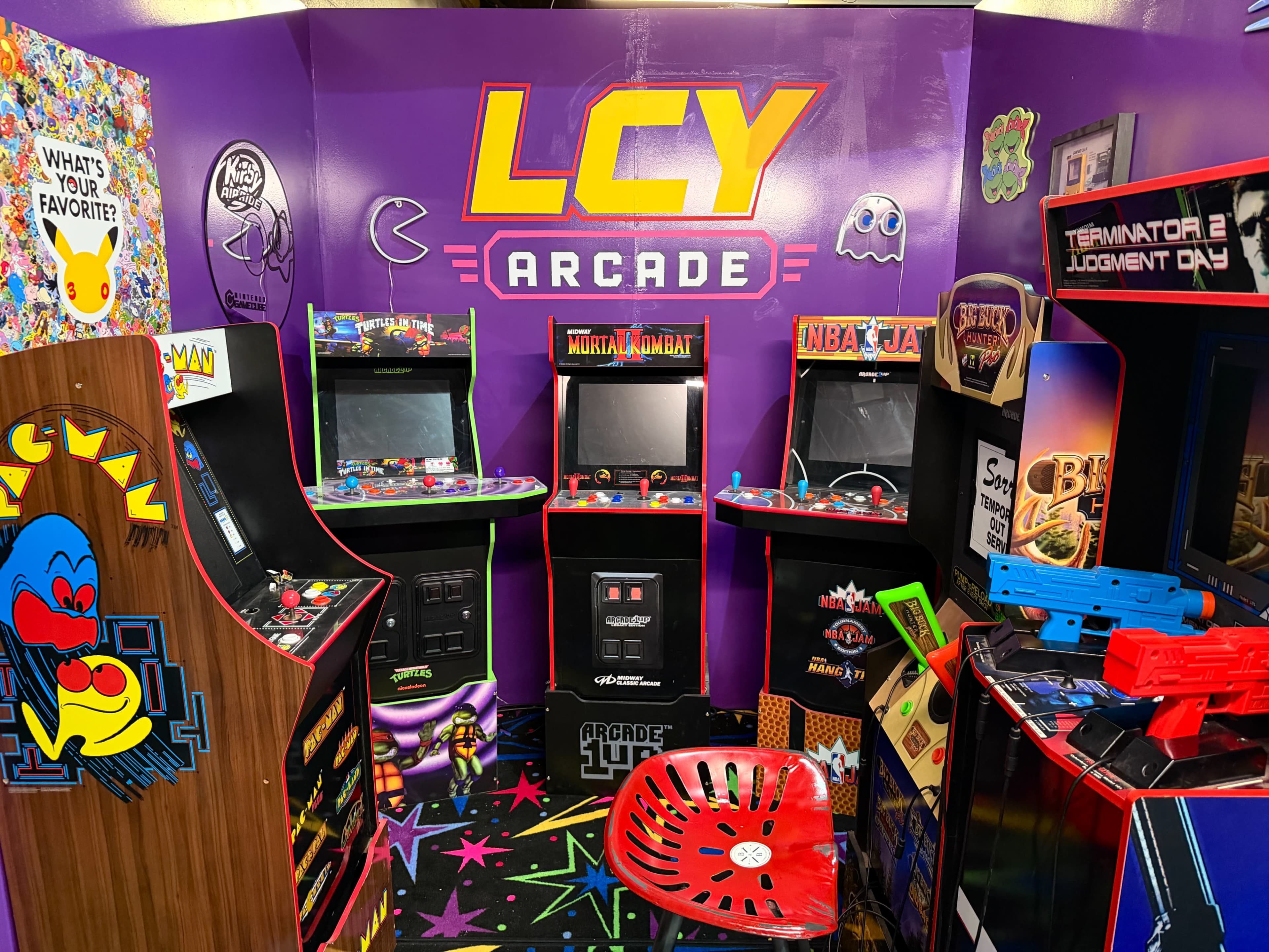 The image depicts a colorful arcade room featuring various retro arcade machines, including titles like Pac-Man and Mortal Kombat, with vibrant wall decorations and a patterned floor.