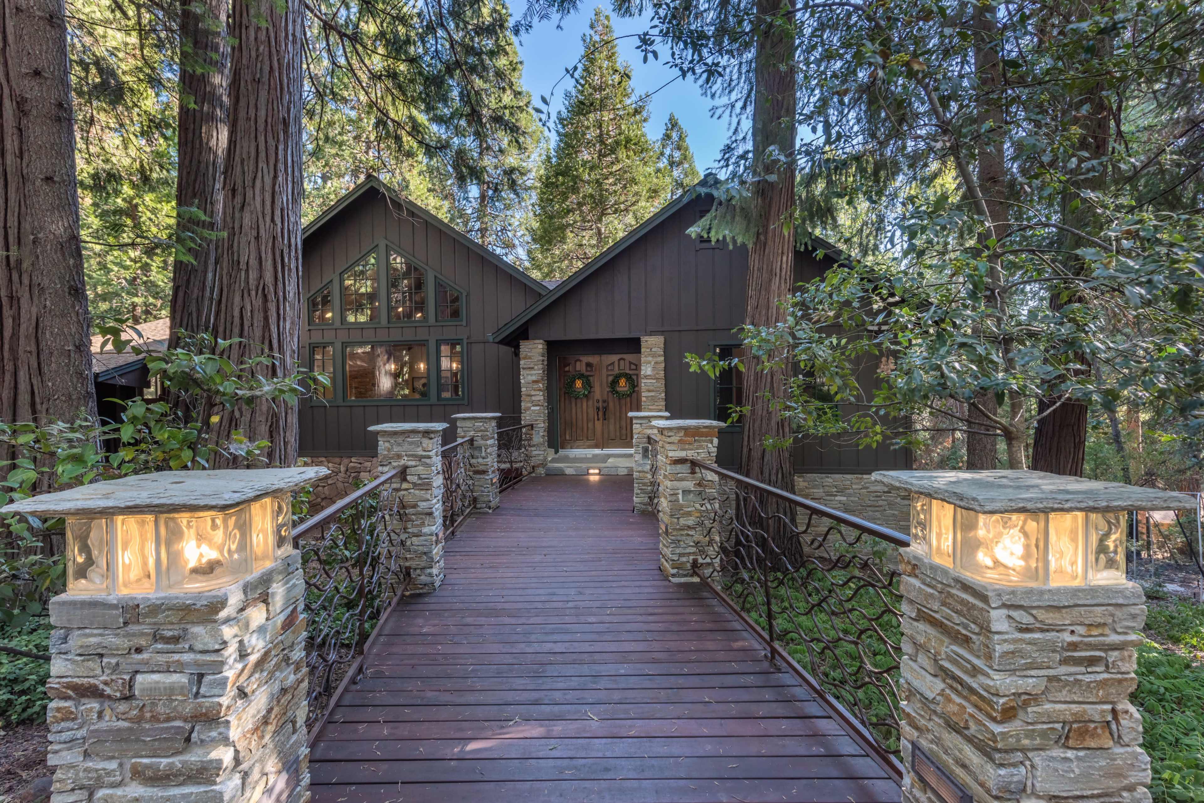 A wooden walkway leads to a dark-colored house surrounded by tall trees, with stone lanterns on either side.