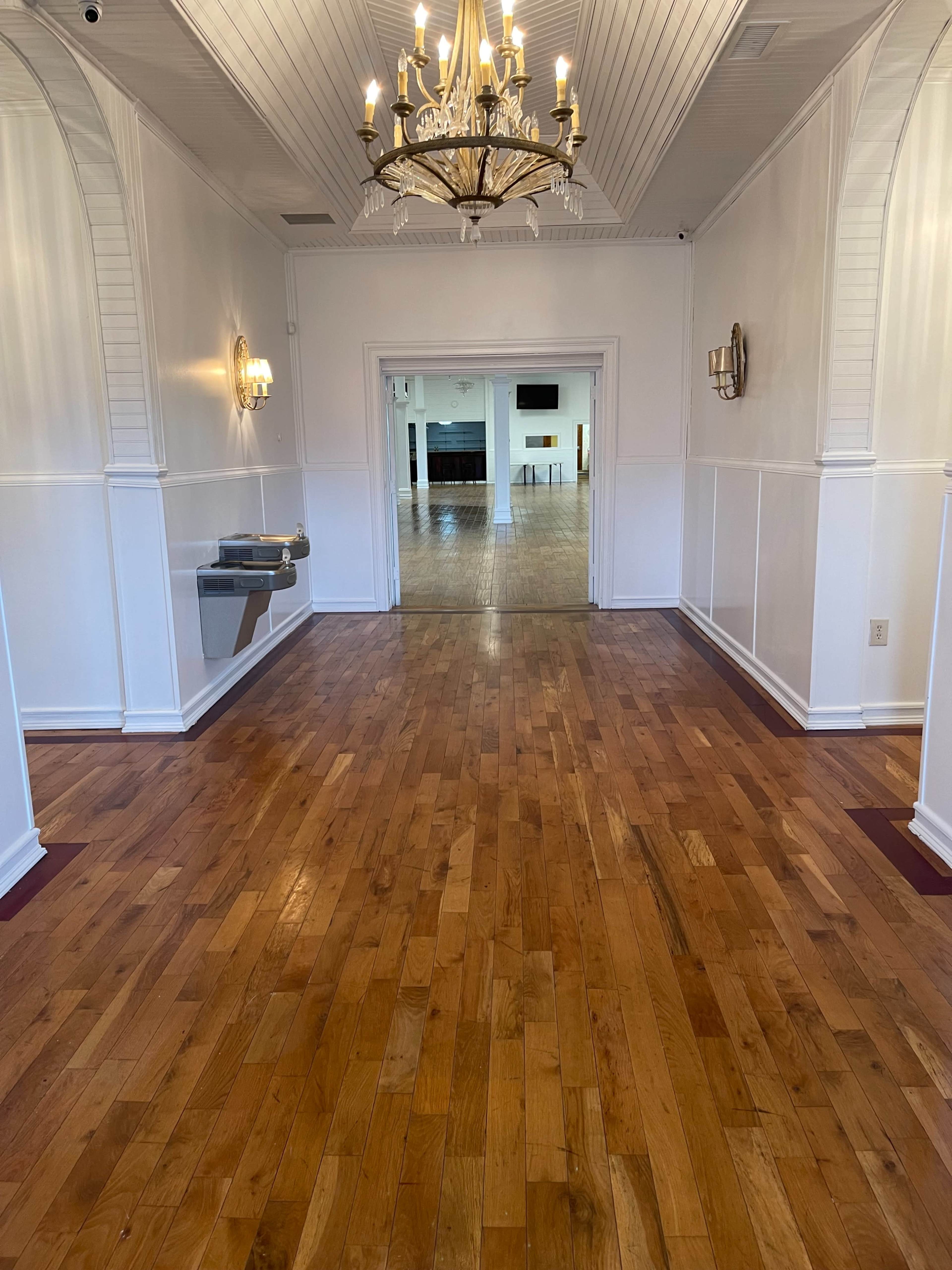 The image shows a well-lit hallway with wooden flooring, leading to a spacious room visible in the background.
