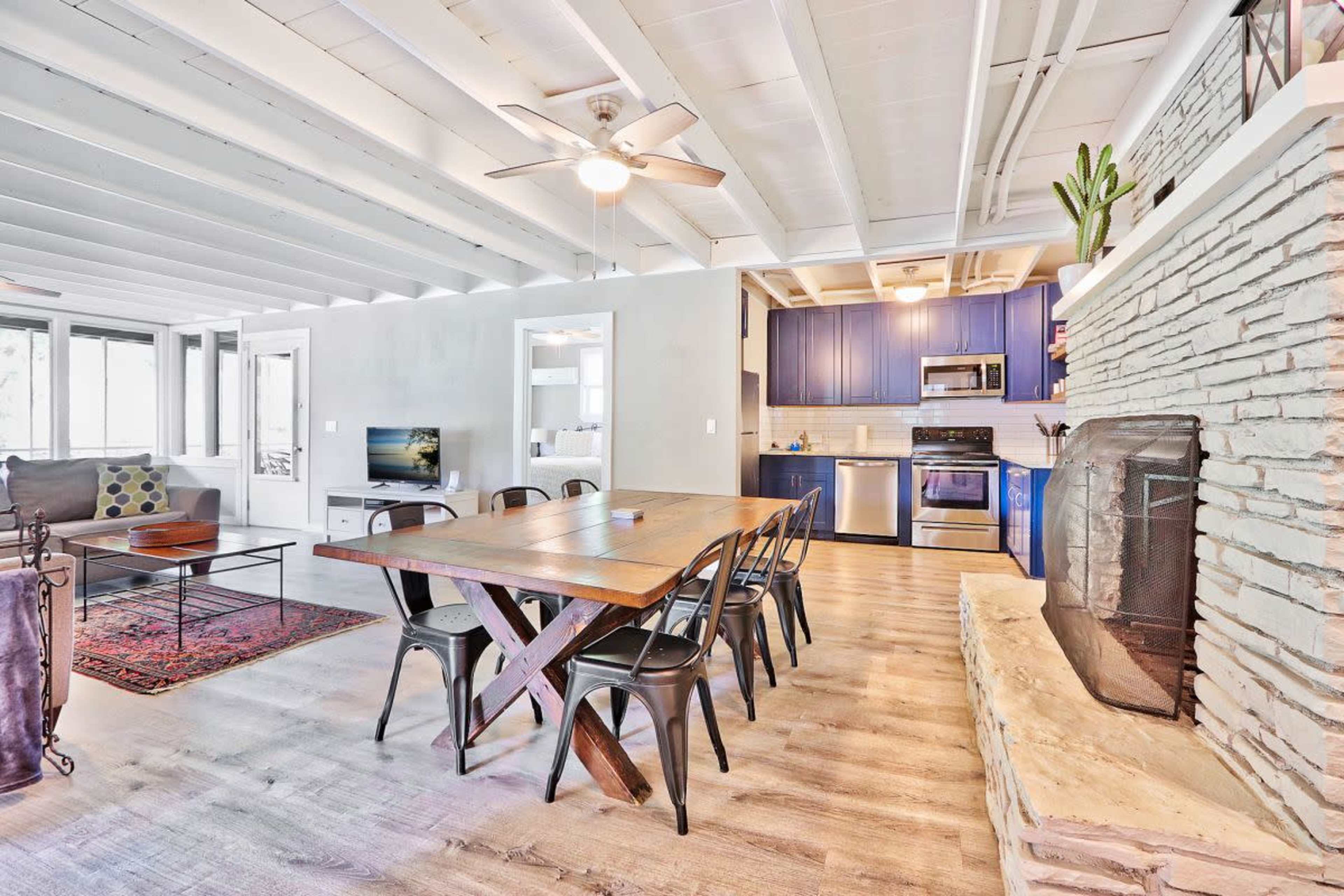 A modern open-concept living and dining area features a wooden table, black chairs, a stone fireplace, and blue kitchen cabinets.