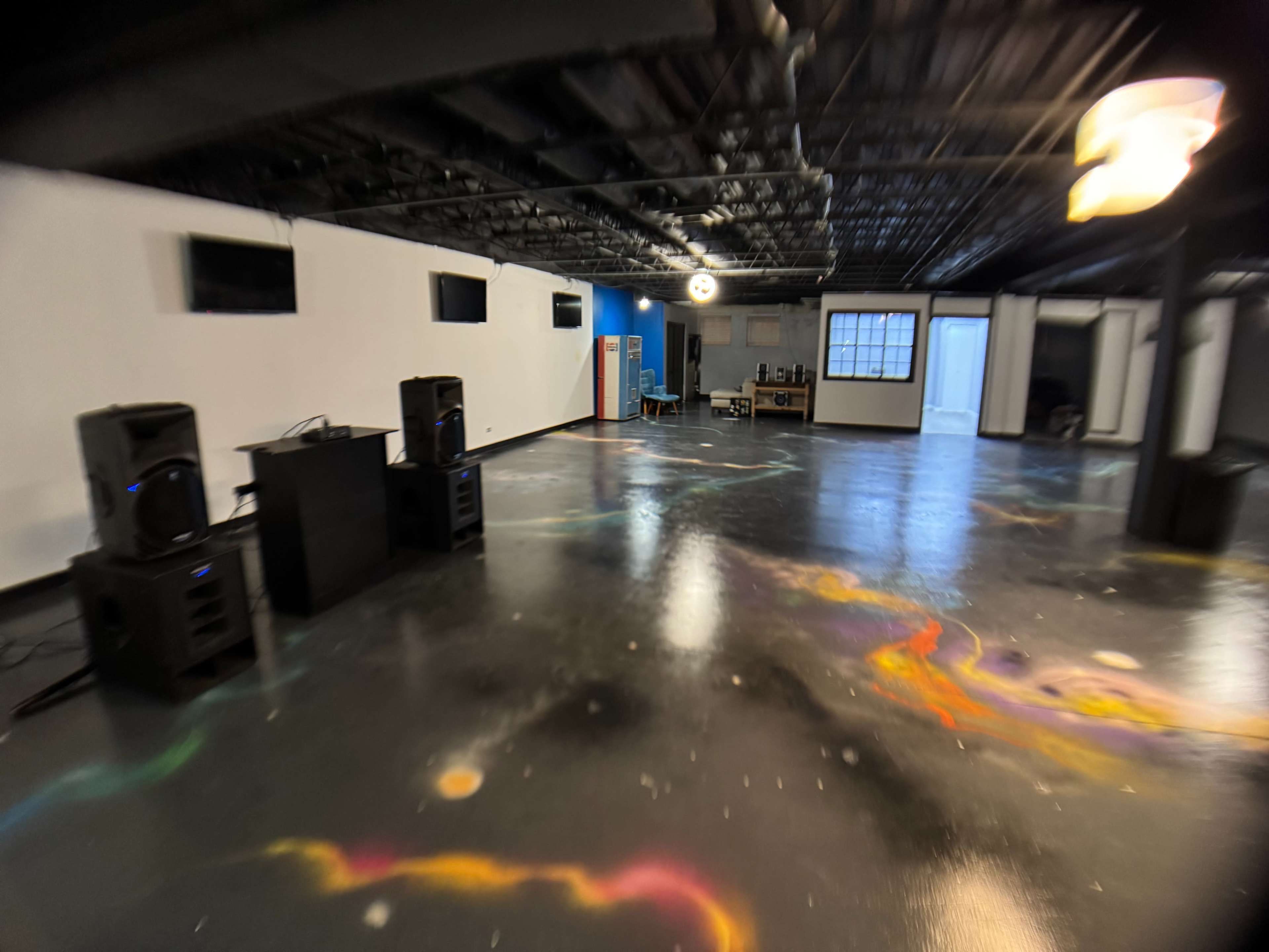 The image shows a spacious room with black floors featuring colorful paint splashes, two large speakers on either side, and several windows and furniture in the background.