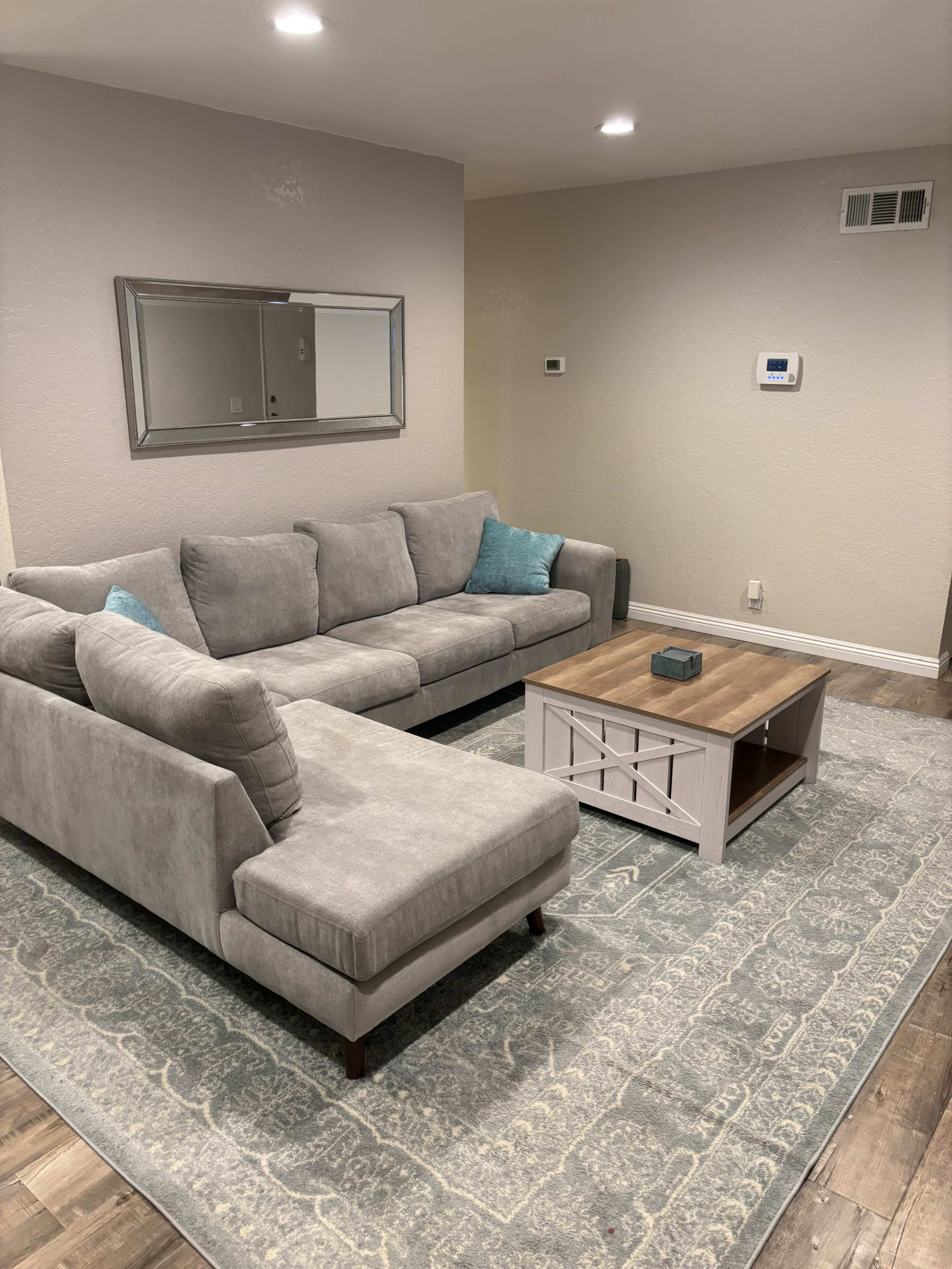 A gray sectional sofa with two accent pillows is positioned beside a wooden coffee table on a patterned area rug in a modern living room.