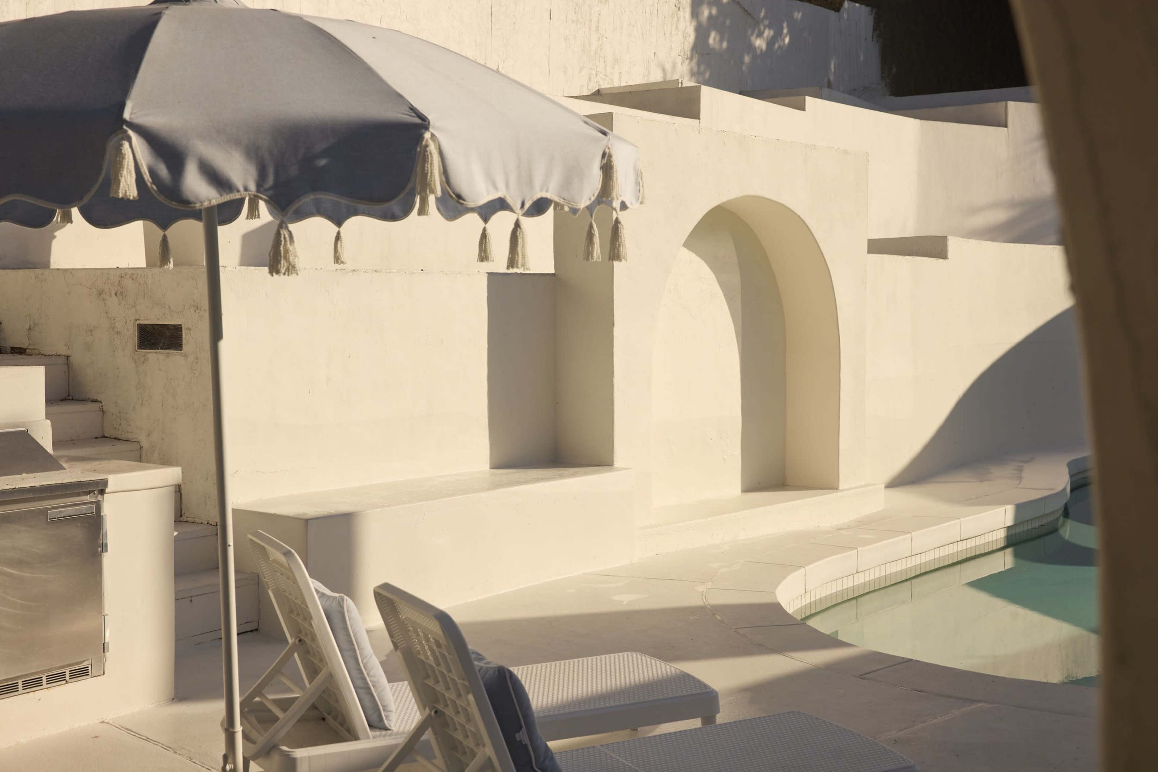 The image shows a serene pool area featuring white walls, a blue umbrella, and two lounge chairs beside a swimming pool.