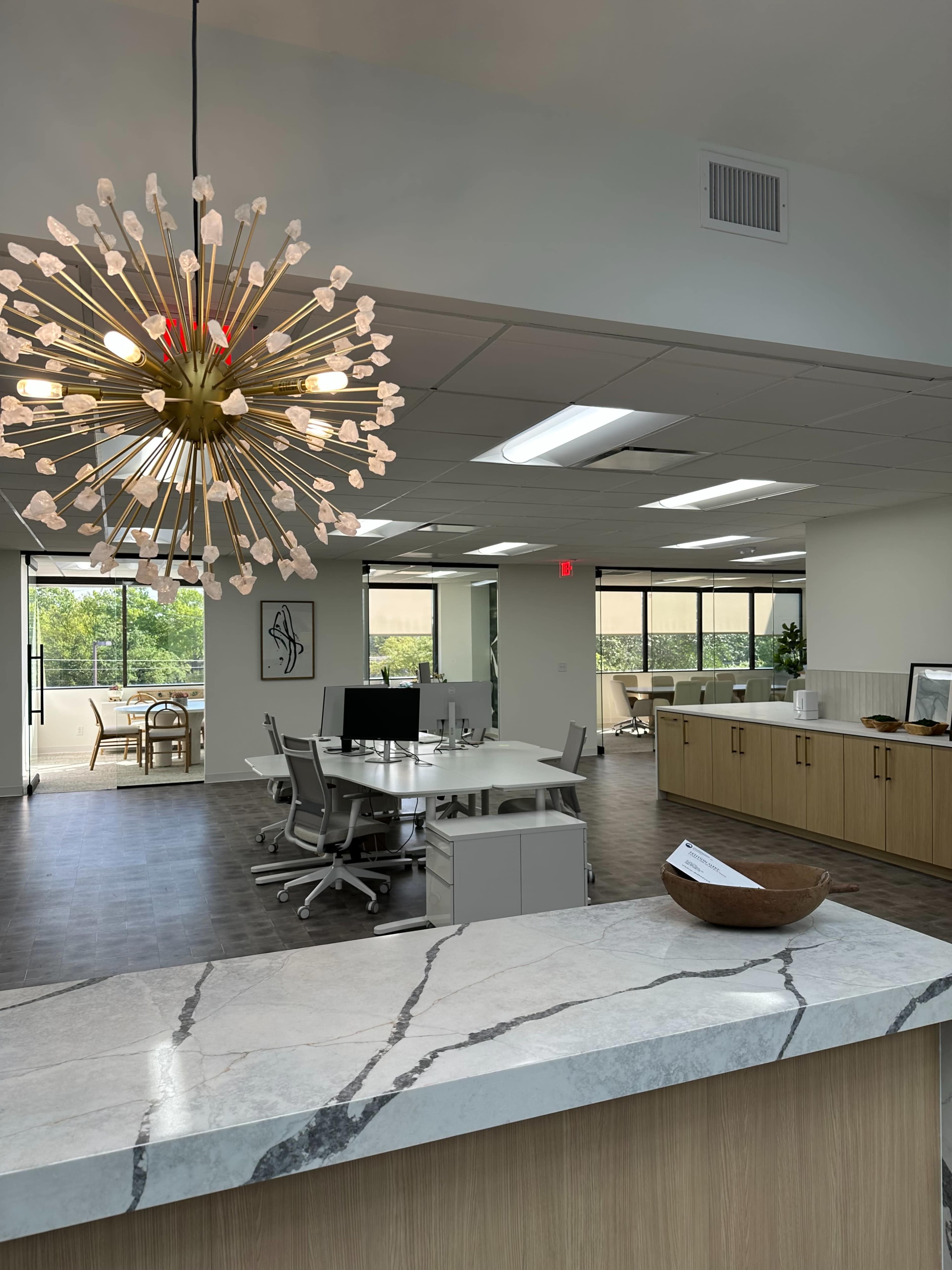 The image shows a modern office space featuring a large light fixture, a reception area with a marble countertop, and workstations in an open layout with large windows.